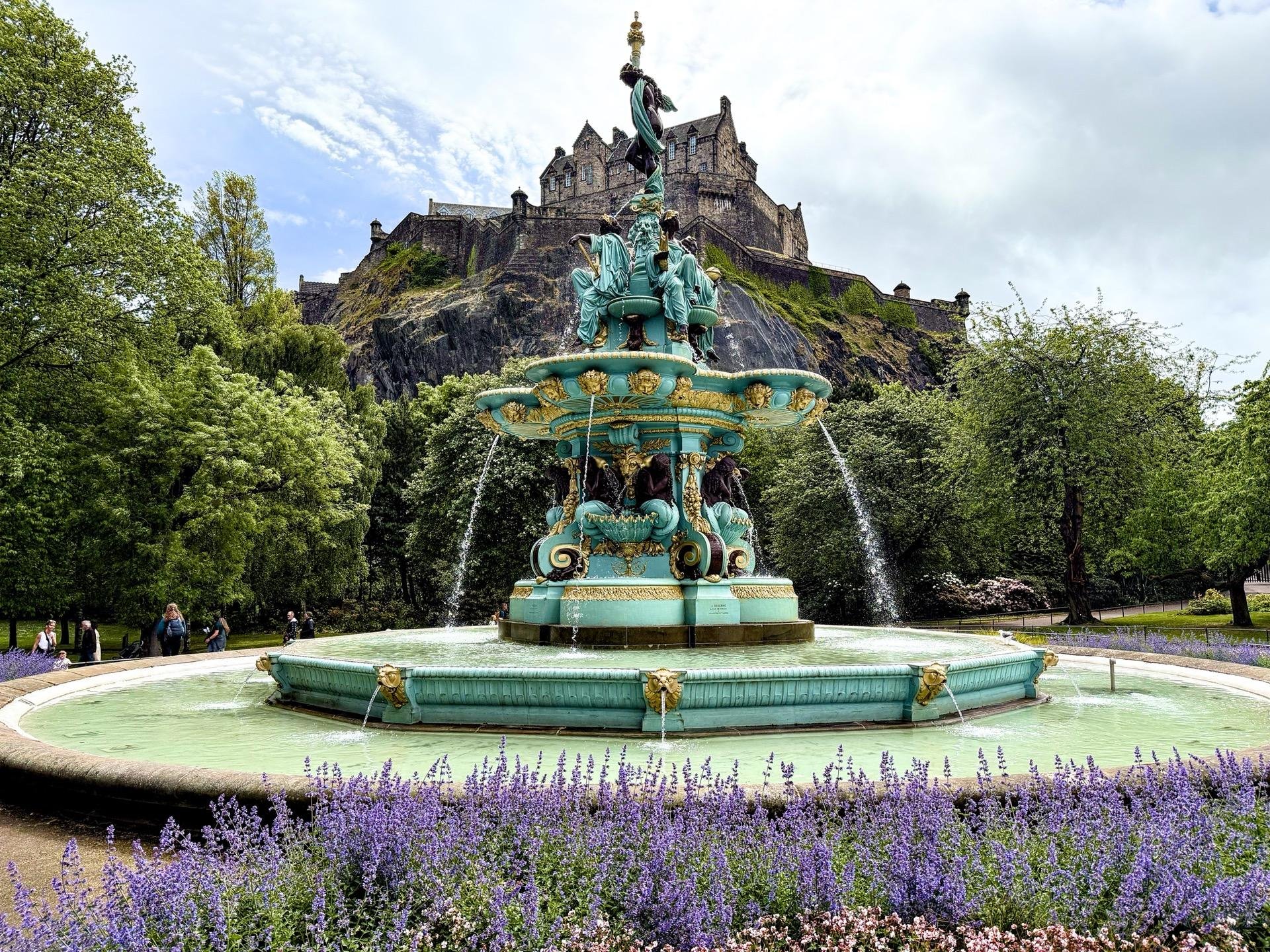 Ornate turquoise fountain with multiple tiers and sculptures, foregrounded by lavender blooms, with a towering castle on a hill in the background under a cloudy sky.