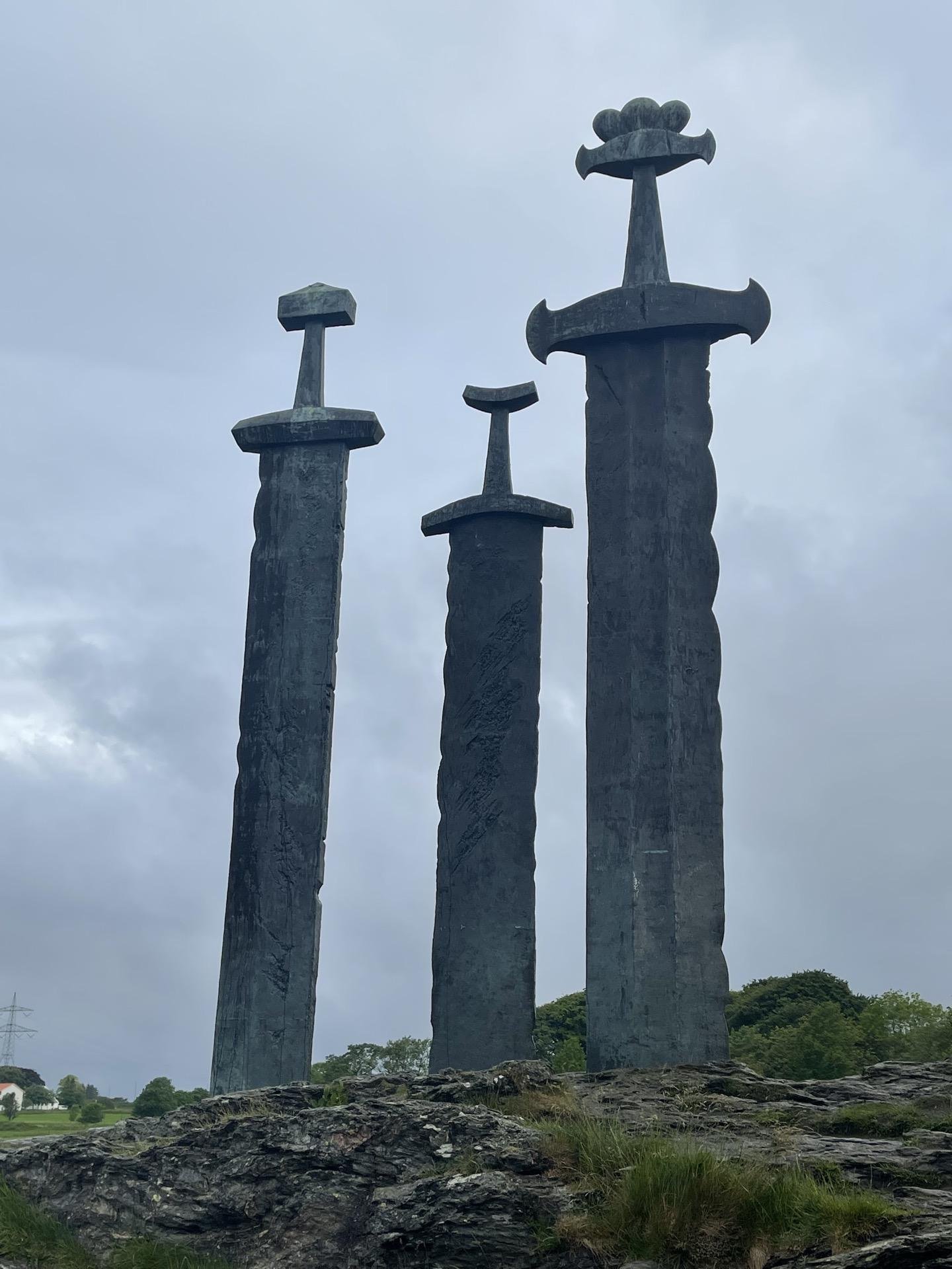 Three large, vertically-standing sword sculptures on a rock base under an overcast sky. The swords differ in size and design, each featuring distinct pommels and cross-guards, sculpted in a rough, textured dark metal. The surrounding landscape includes low grass and distant trees.
