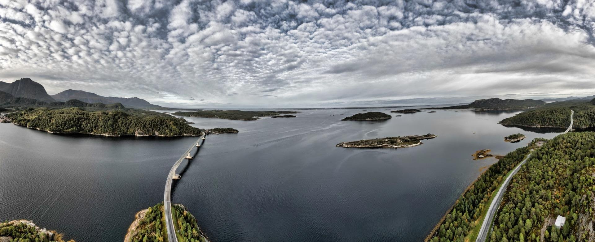 Panoramic view of a curved bridge spanning a calm bay surrounded by forest-covered islands and hills under a cloudy sky. The road extends from the foreground into the distance, flanked by dense green tree lines and scattered islets in the water.