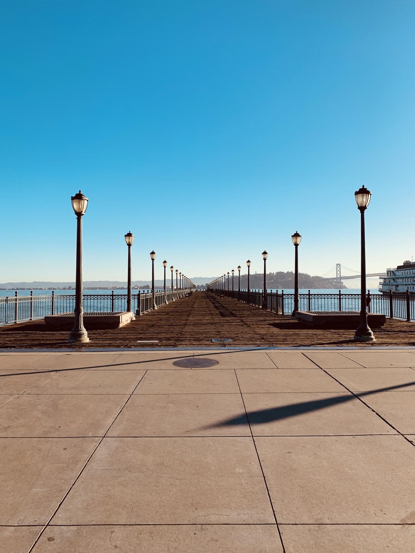 Pier extending into the water with a series of evenly spaced lamp posts on either side, under a clear blue sky. The concrete walkway is divided into large rectangular panels, leading toward a distant structure partially visible. A faint shadow stretches across the ground from the left. The Bay Bridge is visible in the far left background.