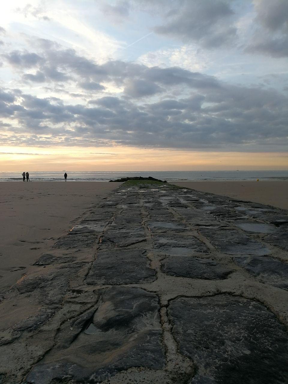 A weathered stone causeway stretches toward the horizon on a sandy beach at sunset. The sky is scattered with clouds and streaked with contrails, illuminated by the fading light. In the distance, several individuals and a dog are visible walking along the shore. The beach appears vast and tranquil under the dimming sky.