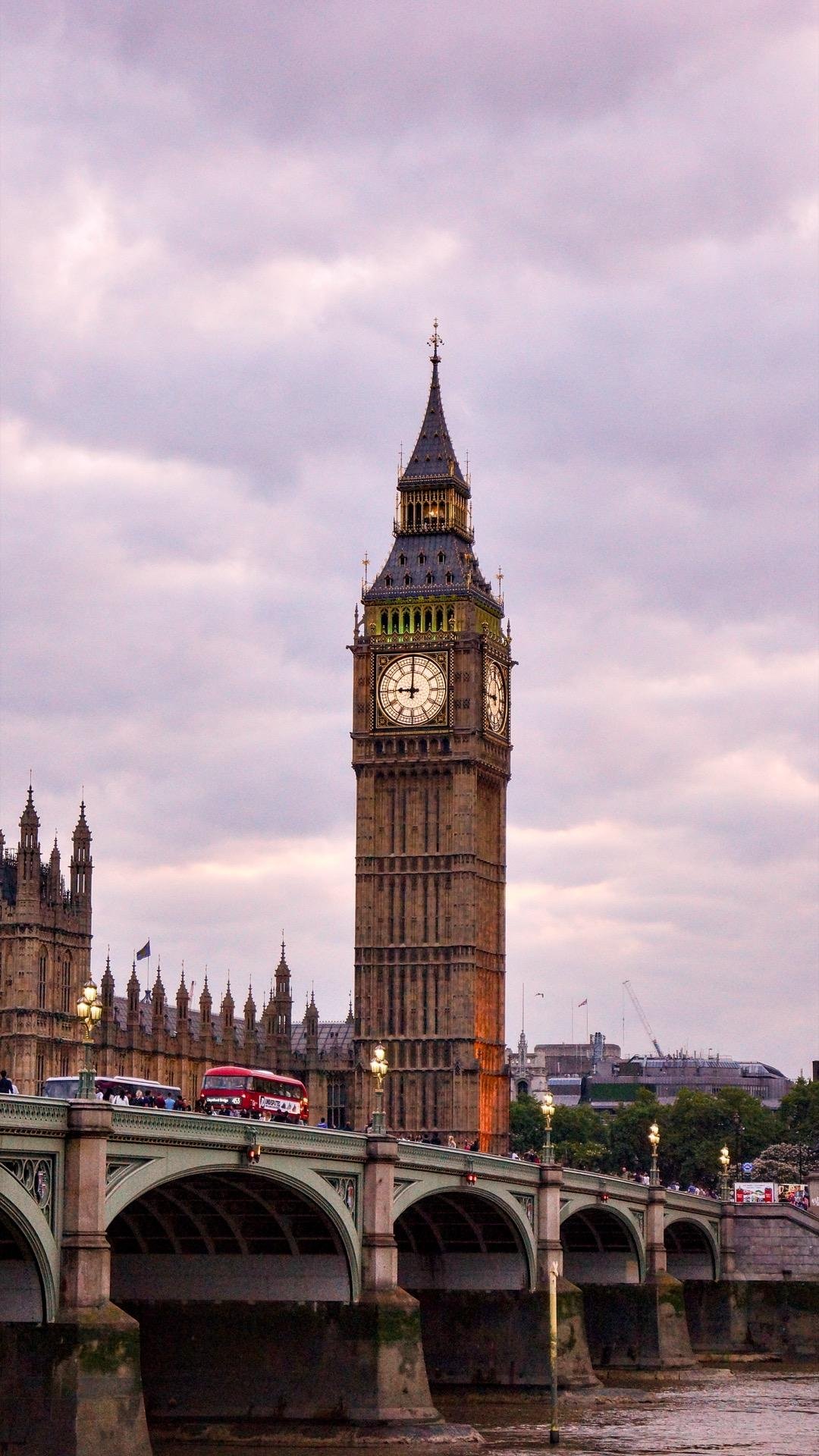 A vertical image of Elizabeth Tower, commonly known as Big Ben, against a cloudy sky during twilight. The tower is illuminated, highlighting its intricate Gothic architecture and the famous clock face. Below, the Westminster Bridge spans over the River Thames with archways and light posts. A red double-decker bus is visible on the bridge, along with several pedestrians.