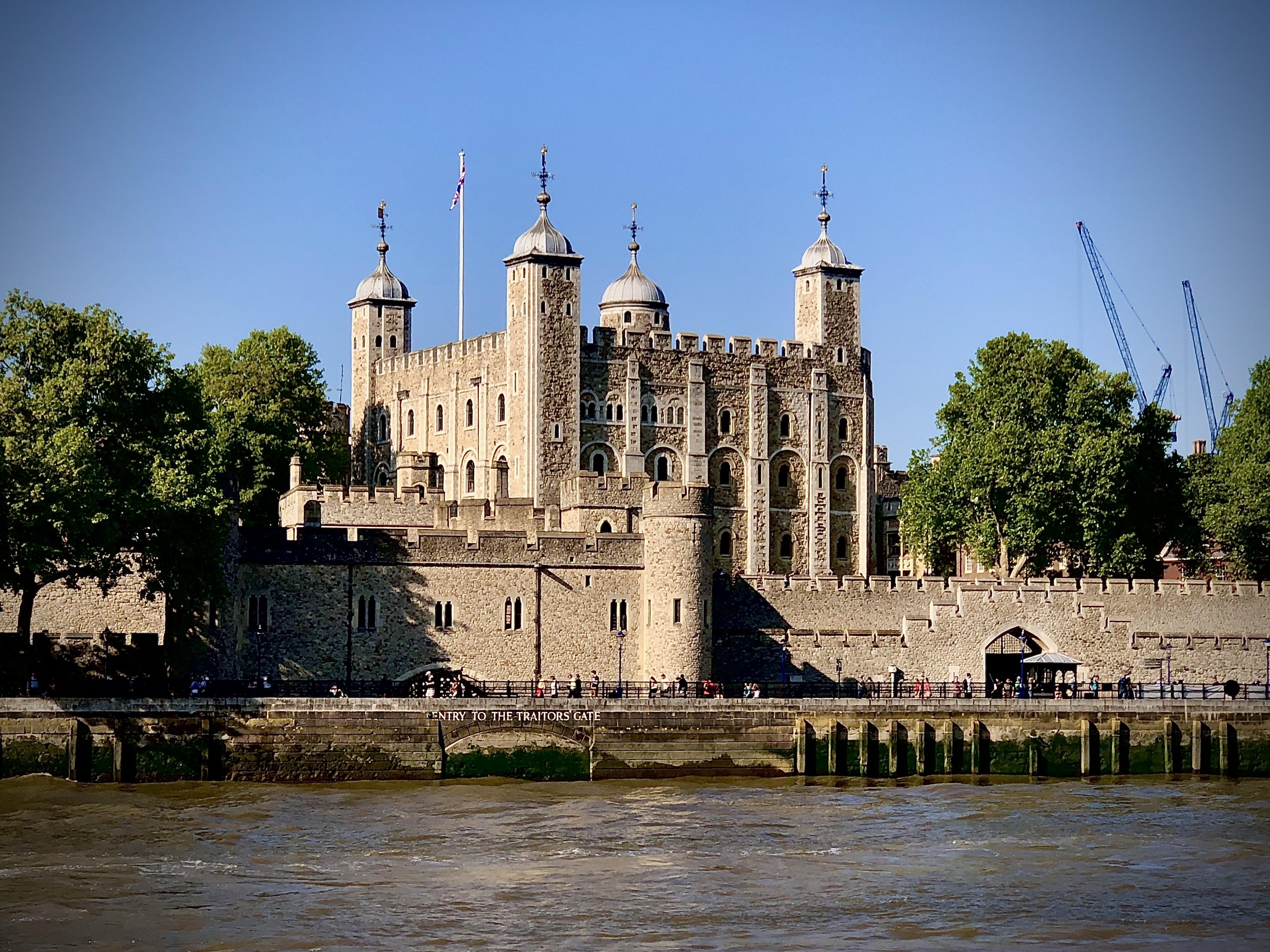 Large, historic stone castle with multiple turrets and a distinctive gate, located beside a river. Several people are visible near the base, and the sky is clear and blue. A small text on a wall reads "Entry to the Traitors' Gate."