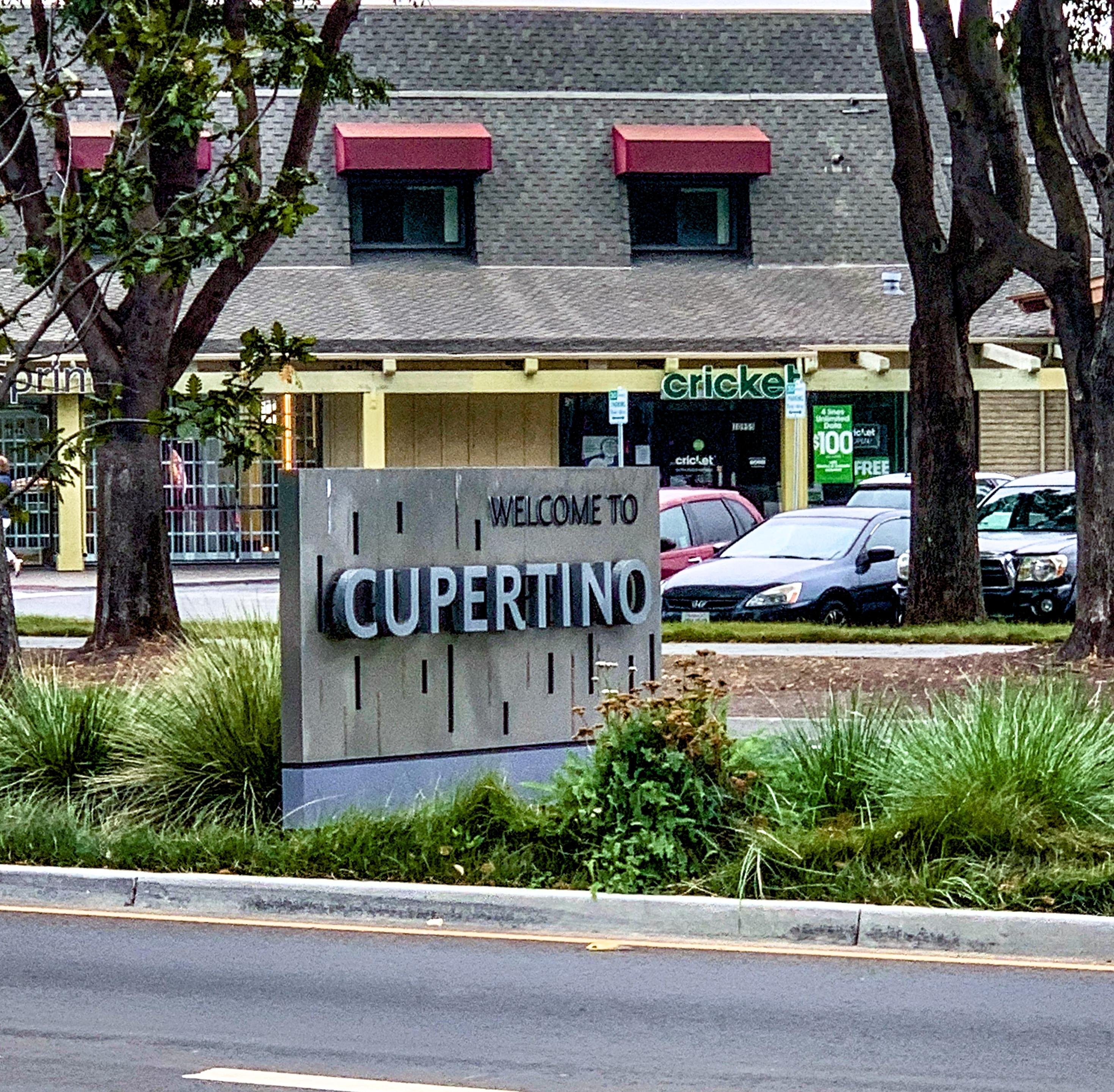Gray sign reading "WELCOME TO CUPERTINO" in bold cut-out letters, positioned in front of a landscaped area along a street. In the background, a two-story commercial building with a green Cricket Wireless store and parked cars are visible.