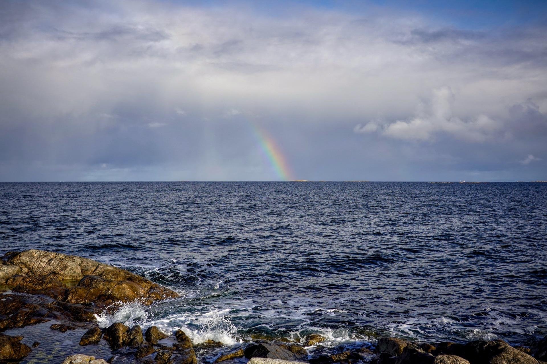 A seascape featuring choppy, blue waters and a rocky shoreline in the foreground, under a partly cloudy sky with a faint rainbow visible in the distance. Waves gently break against the rocks.