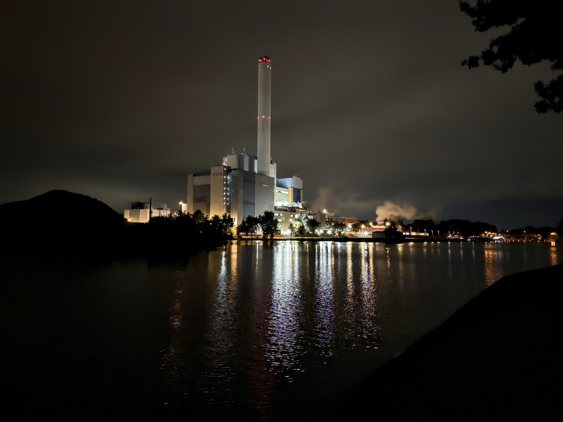 Nighttime view of an illuminated industrial plant by a calm water body, with reflections of the lights visible on the water's surface. The plant features multiple buildings and tall chimneys, one prominently displaying red lights at the top. A dark outline of a hill is visible on the left, and a hint of vegetation on the right edge under a cloudy sky.