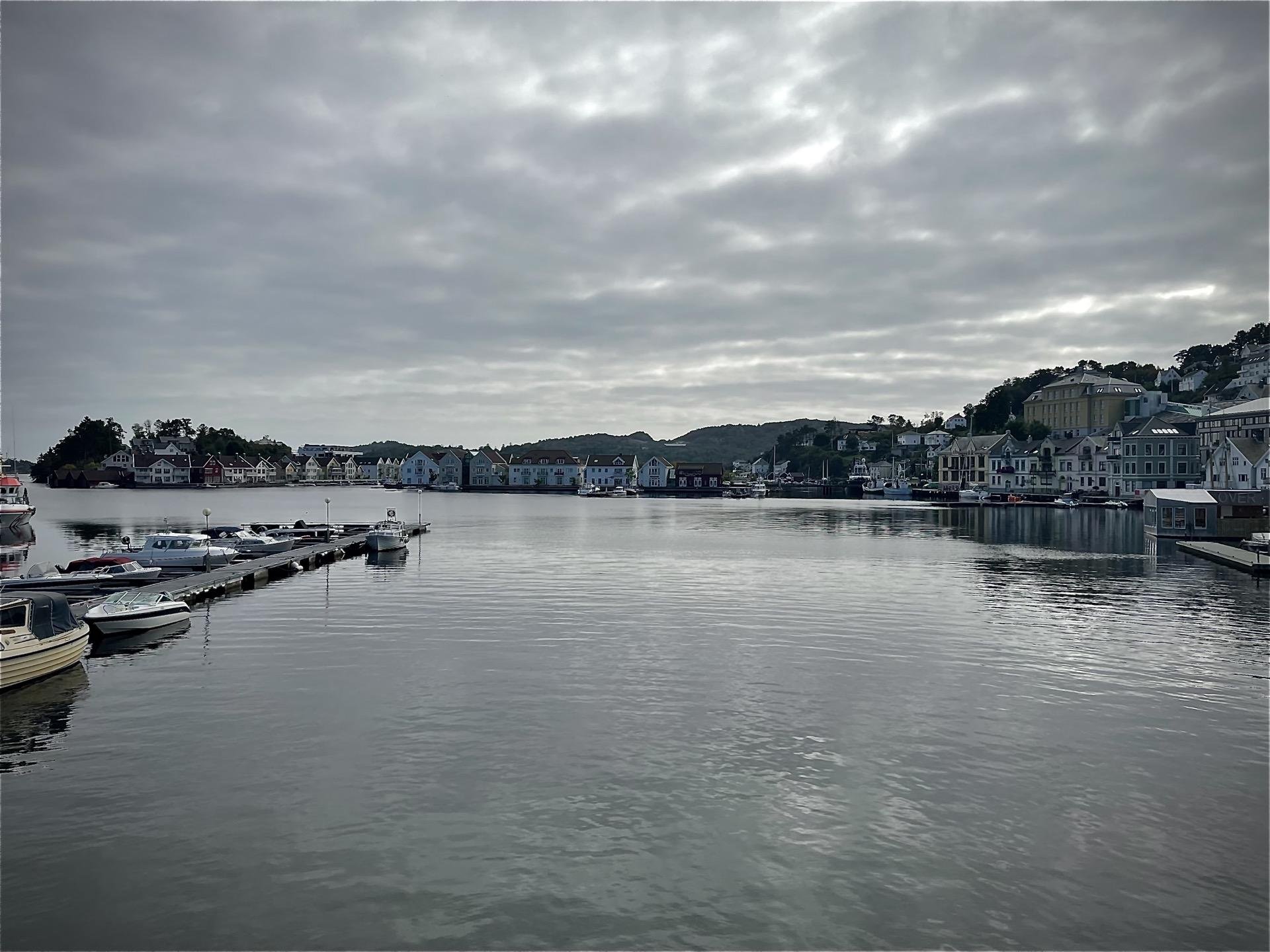 Overcast sky above a calm harbor with multiple boats docked on a jetty extending into the water. Colorful houses line the waterfront against a backdrop of rolling hills.