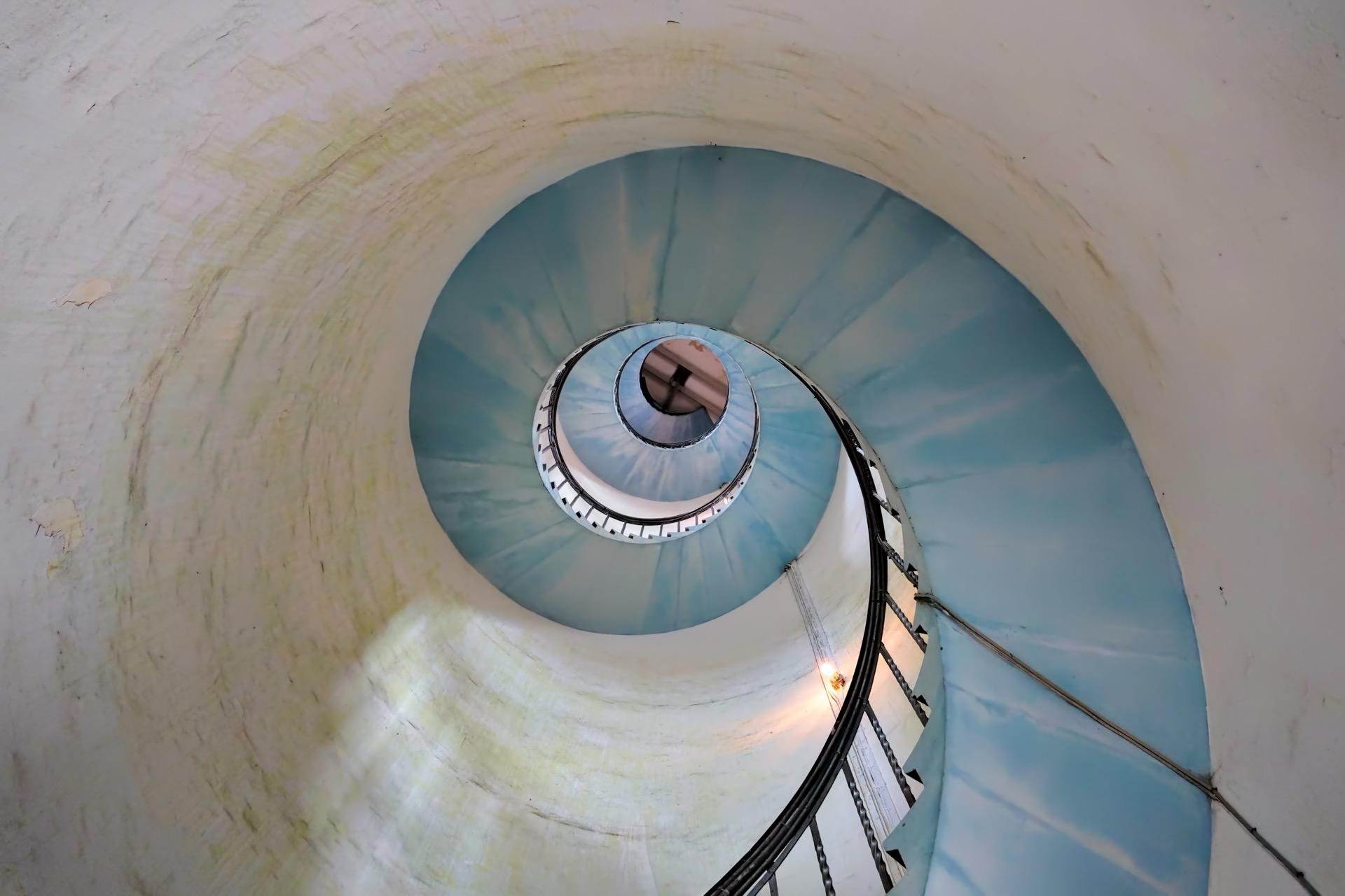 Spiral staircase viewed from below, with a gradually ascending metal railing and steps, lit by a warm light. The stairs create a swirling pattern against the white, round walls marked by subtle green and brown aging. Light filters in softly, casting shadows and giving a dreamy effect to the pale blue center of the spiral.