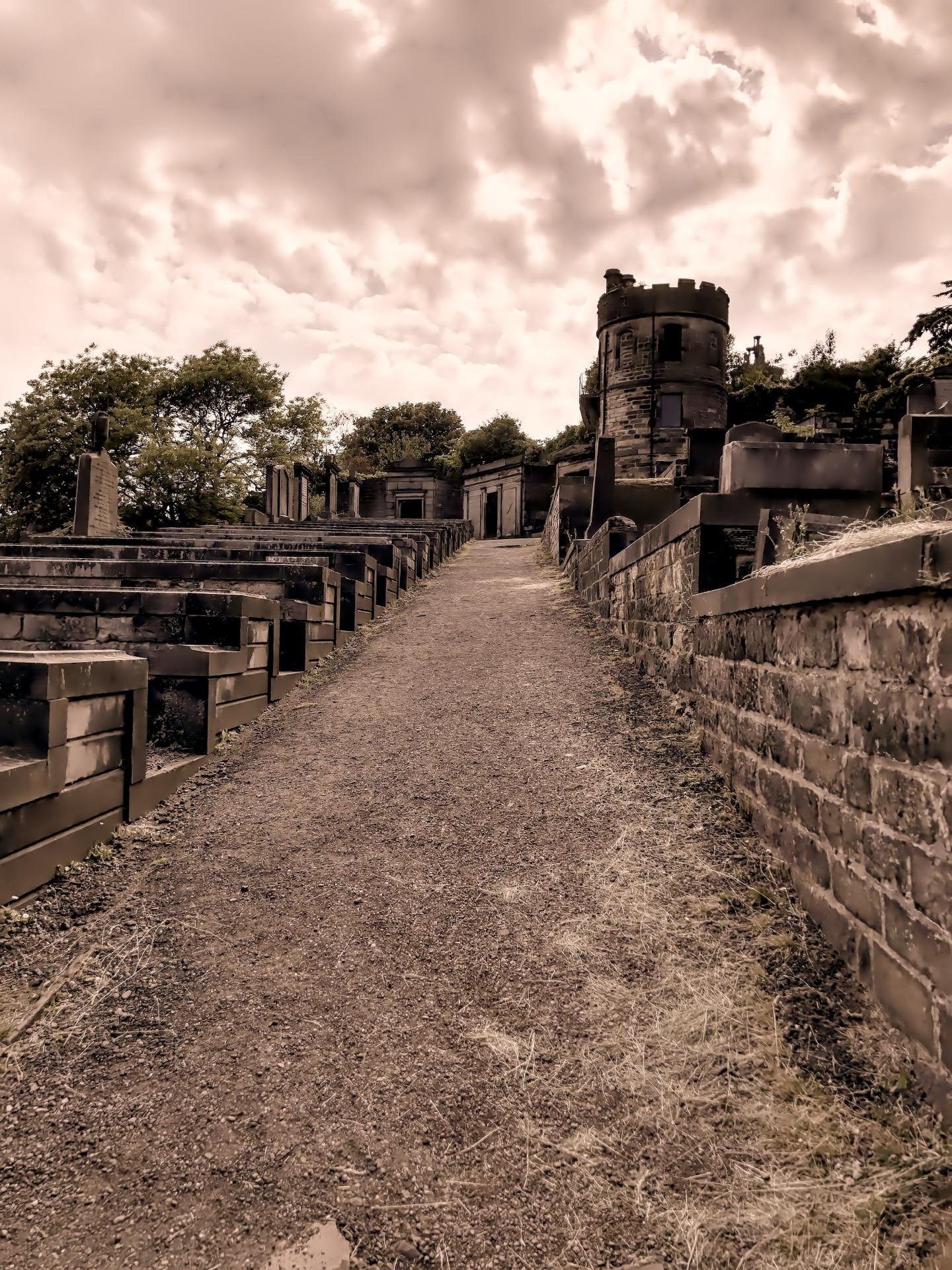 Narrow dirt pathway flanked by aged, deteriorating crypts and tombstones under a cloudy sky in a historic cemetery. The path is bordered by stone structures and leafy trees, leading towards a round, tower-like stone edifice on the right. The scene is cast in a sepia tone, enhancing the somber, antique atmosphere of the setting.