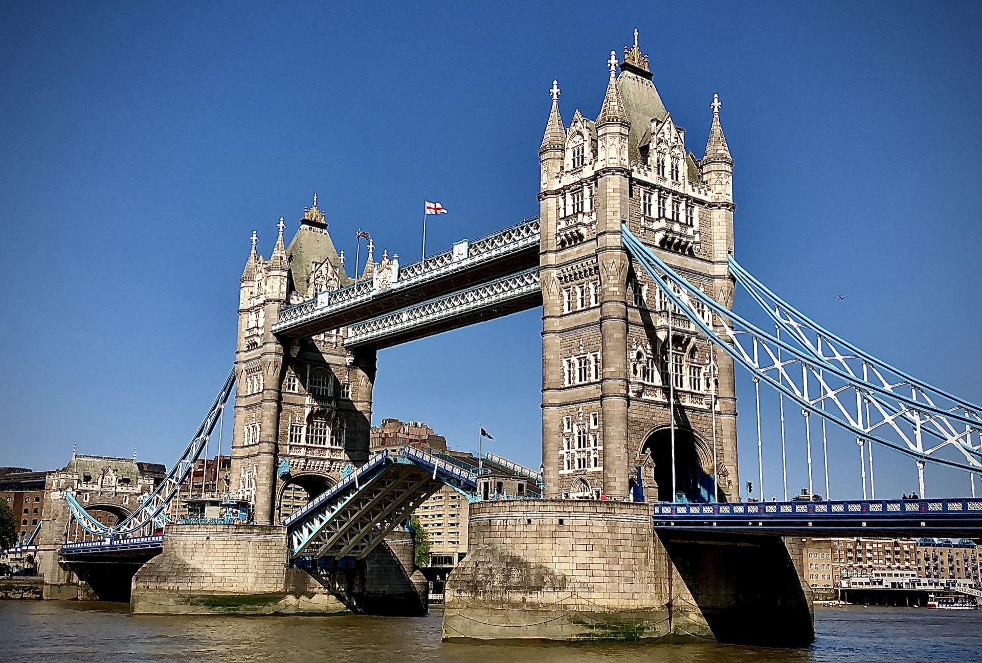 Tower Bridge in London, featuring two prominent towers with pointed roofs connected by upper walkways, set against a clear blue sky. The bridge is partly raised, displaying its bascule (drawbridge) mechanism in action. Visible are intricate architectural details on the Victorian Gothic towers and the blue and white color scheme of the bridge structure. A UK flag flutters atop one tower. The Thames River and riverbanks are visible, with architectural buildings in the background.