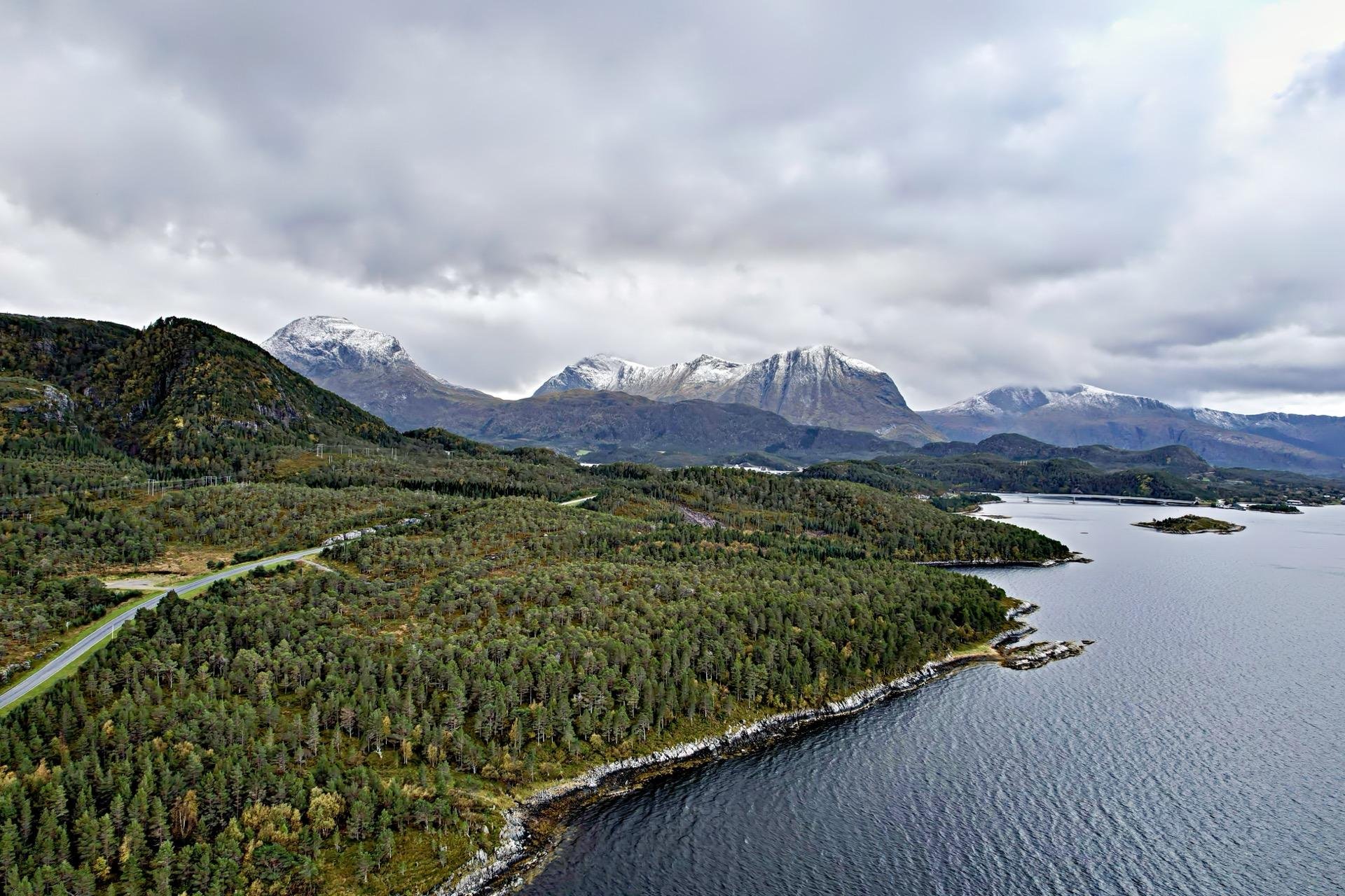 Overhead view of a rugged coastline with a narrow road winding through a dense, green forest, leading towards distant snow-capped mountains under a cloudy sky. The forest is bordered by a calm body of water, reflecting a gray overcast, and the terrain is dotted with small patches of barren land and a few scattered lakes.