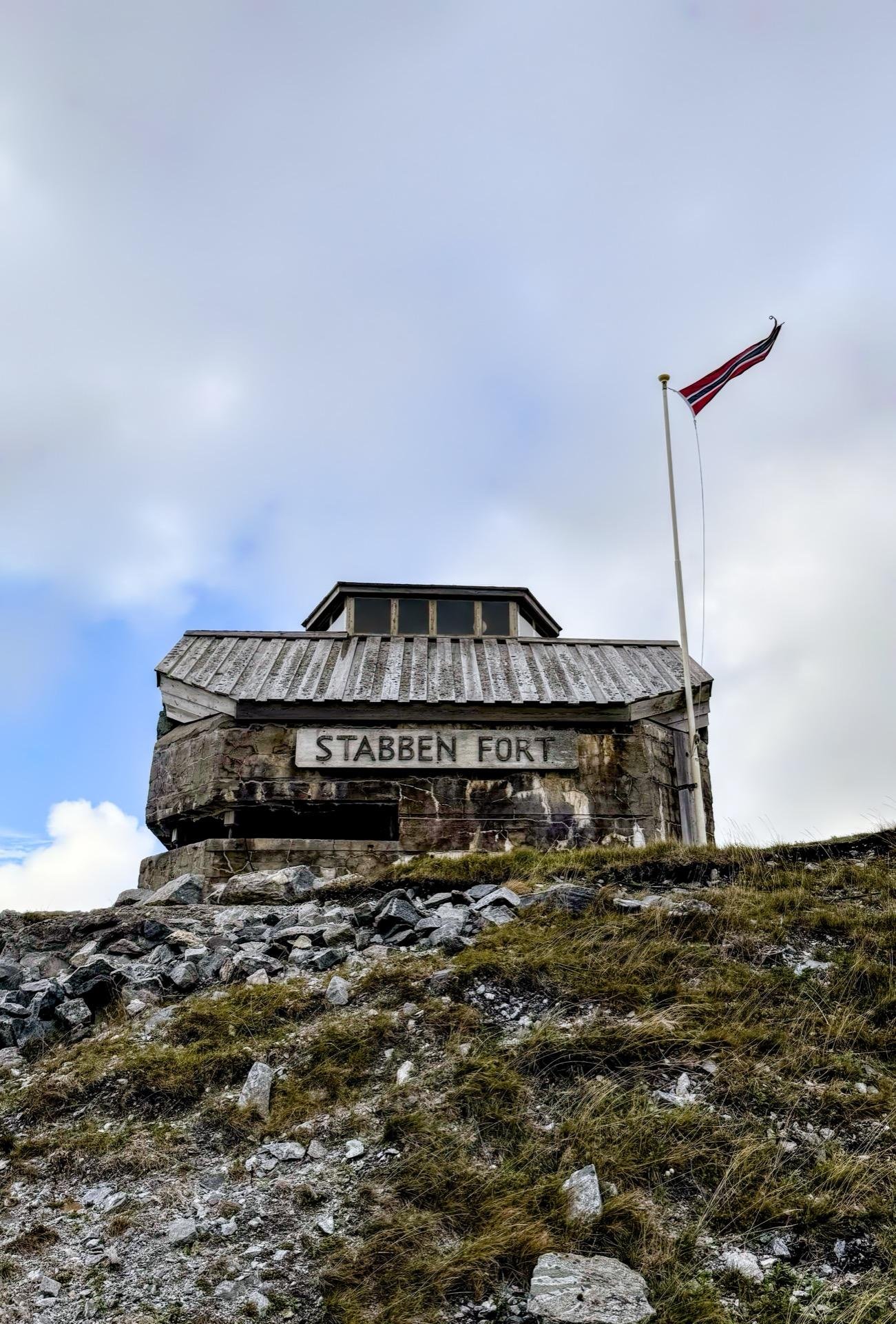 Stone building labeled "Stabben Fort" atop a grassy hill under a cloudy sky, with a Norwegian flag on a pole fluttering to the right. The fort features a rugged base of large stones and a weathered wooden upper structure with a peaked roof.