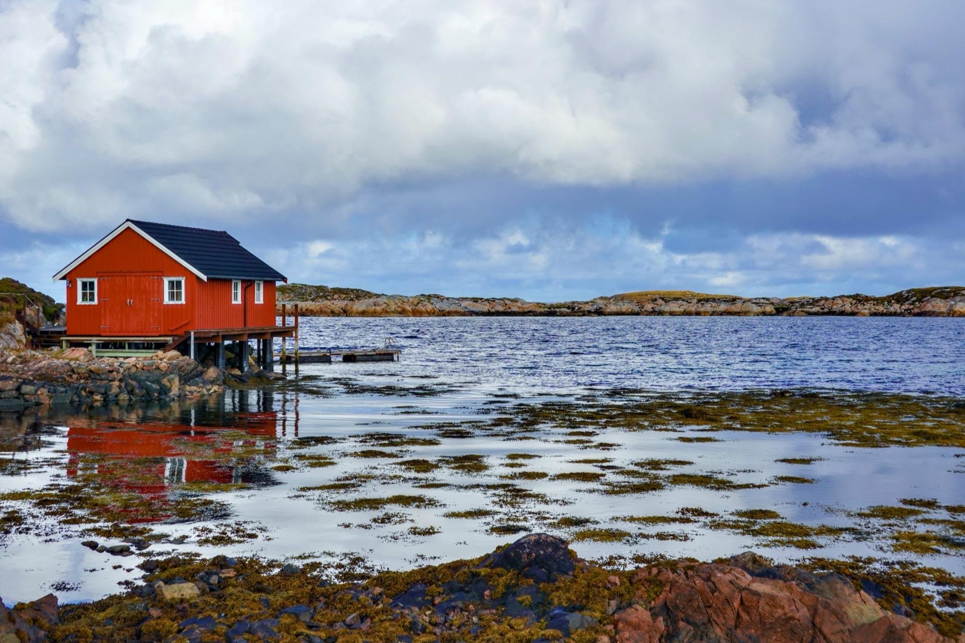 Red wooden house on stilts located over seaweed-strewn water along a rocky coastline, with a backdrop of cloudy skies and distant rocky islets.