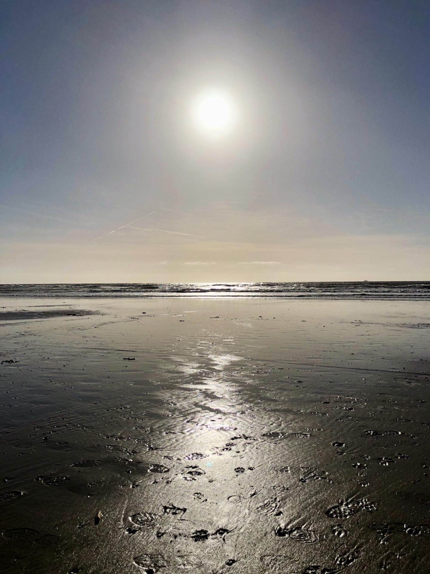 Bright sun hanging directly above the horizon casts a dazzling reflection across a wet sandy beach. The surface of the sand is textured with small footprints and ripples. There are subtle plane trails in the clear blue sky.