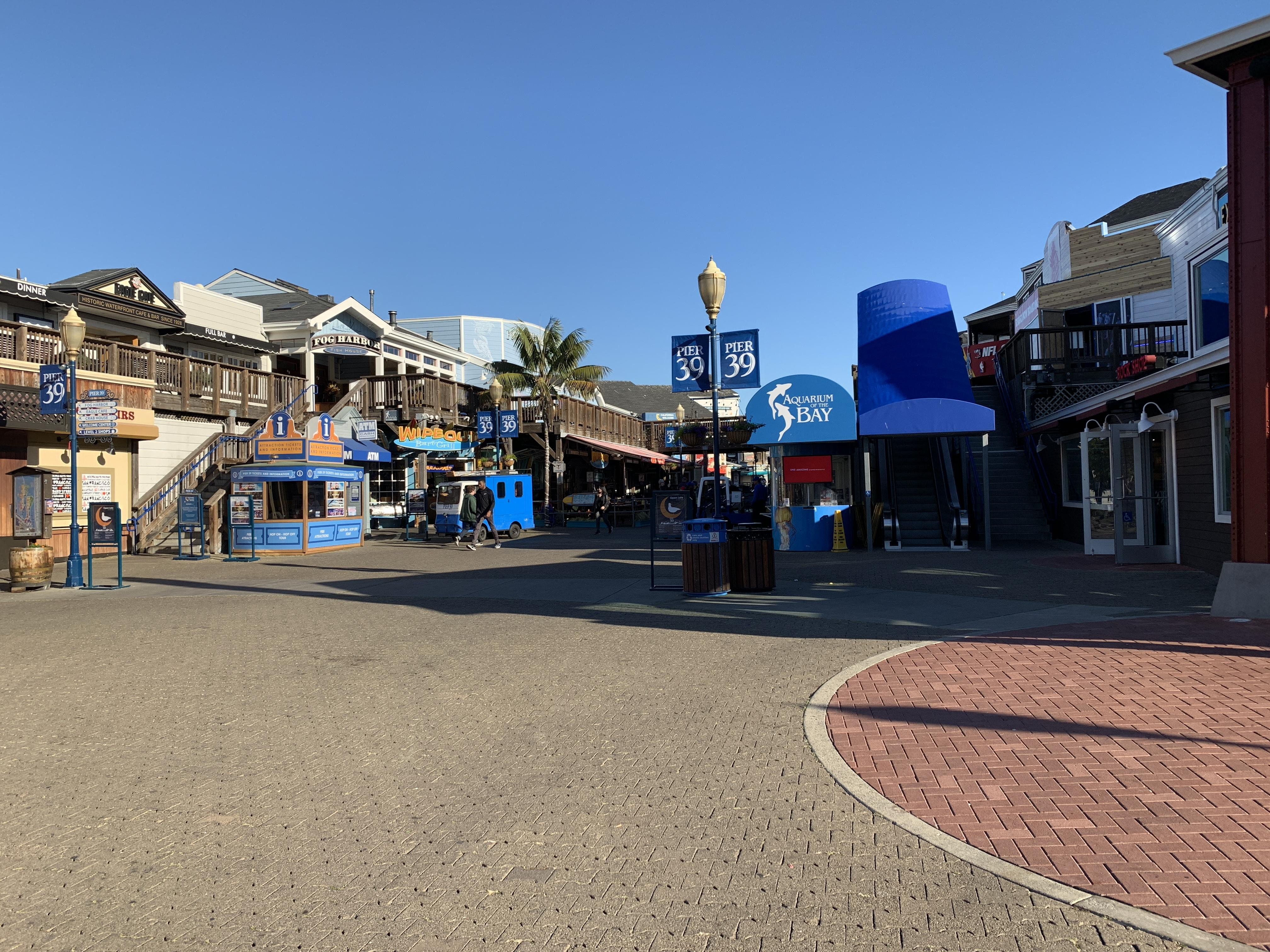 Clear blue sky over a bustling outdoor shopping area at Pier 39, featuring multiple two-story wooden buildings with variety of shops and blue signs. A significant blue circular structure marked “Aquarium of the Bay” stands on the right, and various informational signs and trash bins populate the brick-paved foreground.