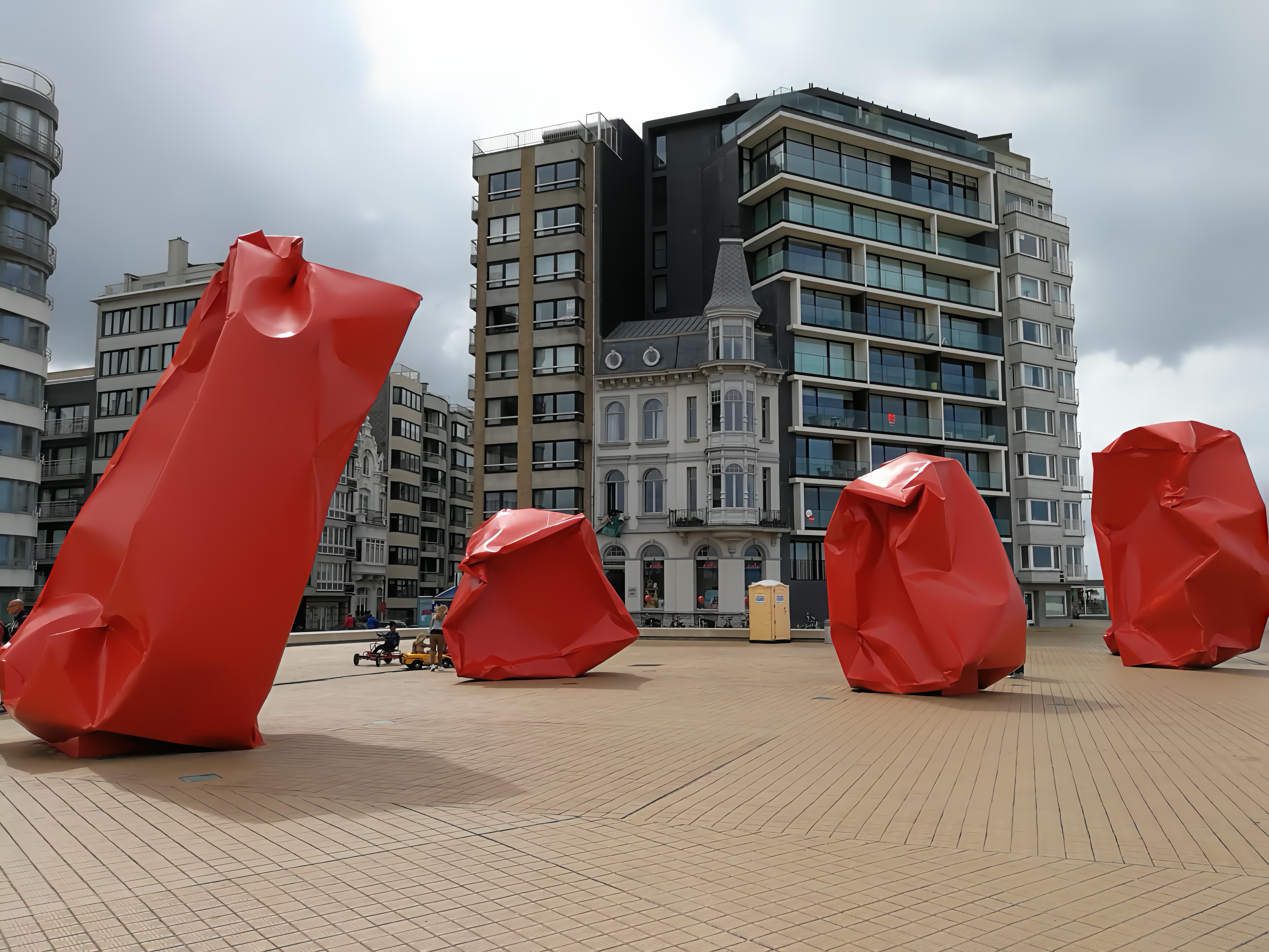 Four large red sculptures resembling crumpled pieces of paper are placed on a spacious, wooden-planked square. The background features a mix of modern and classic architecture, with buildings of varying styles and heights. Overcast skies above suggest a cloudy day. A few pedestrians are visible near the sculptures, providing a sense of scale.
