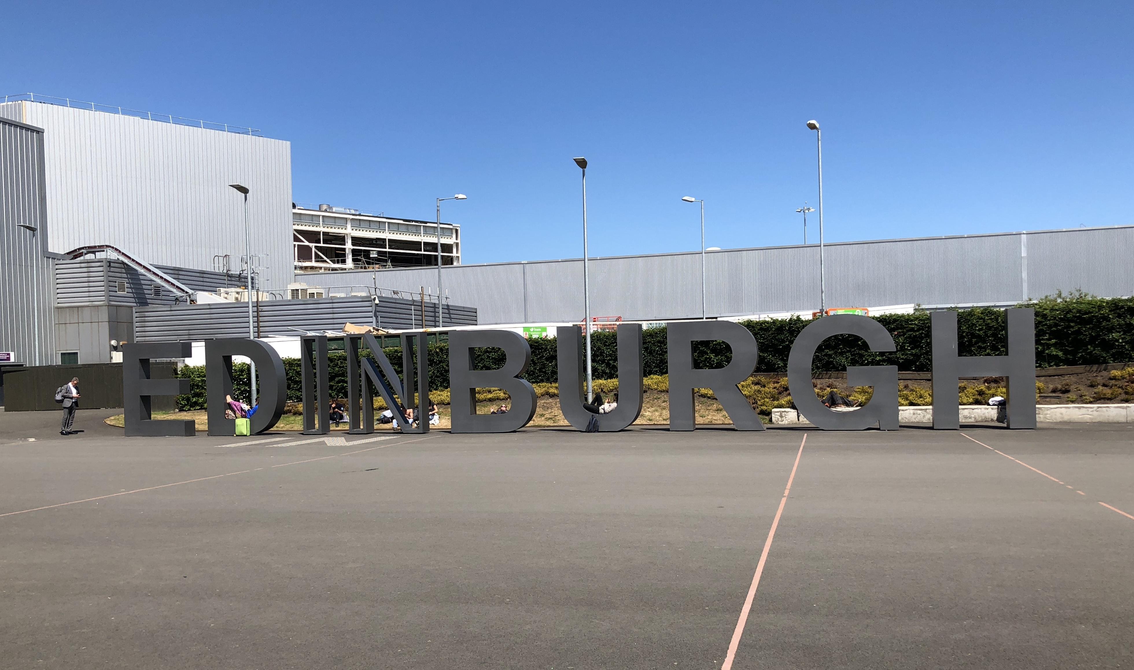 Large freestanding letters spelling "EDINBURGH" installed in a parking area, with a person walking past and another person sitting on one of the letters. Background features a modern building and a partly visible multi-story parking structure under a clear blue sky.