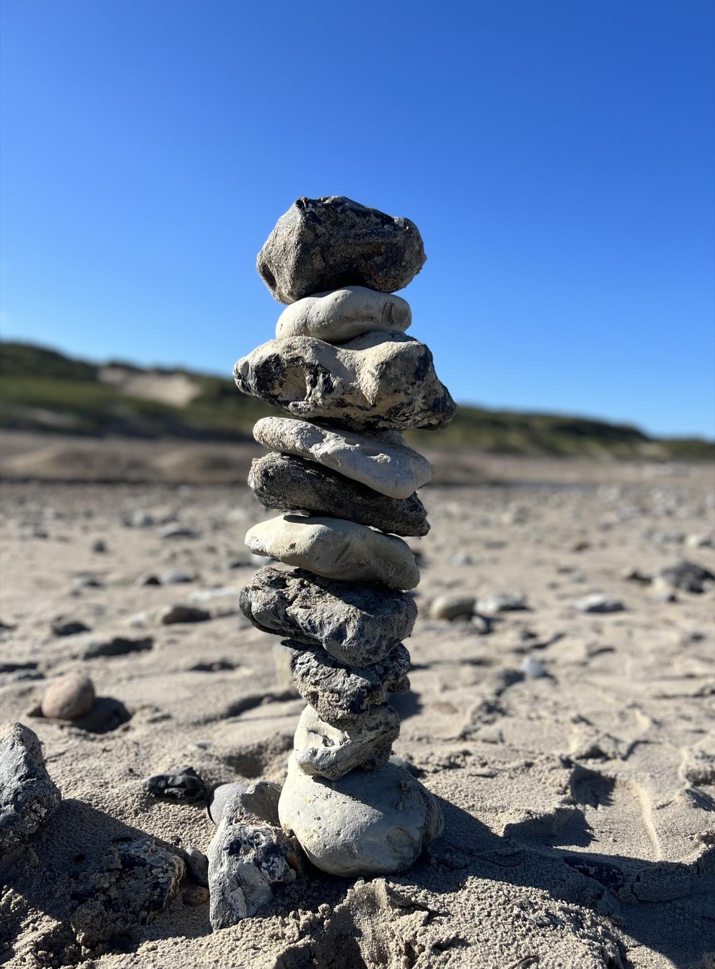 A vertical stack of varied shapes and sizes of rocks balanced precariously on a sandy beach. Each rock has different textures and colors, primarily shades of gray and white. The background is softly blurred, showing a clear blue sky and a faint outline of sand dunes.