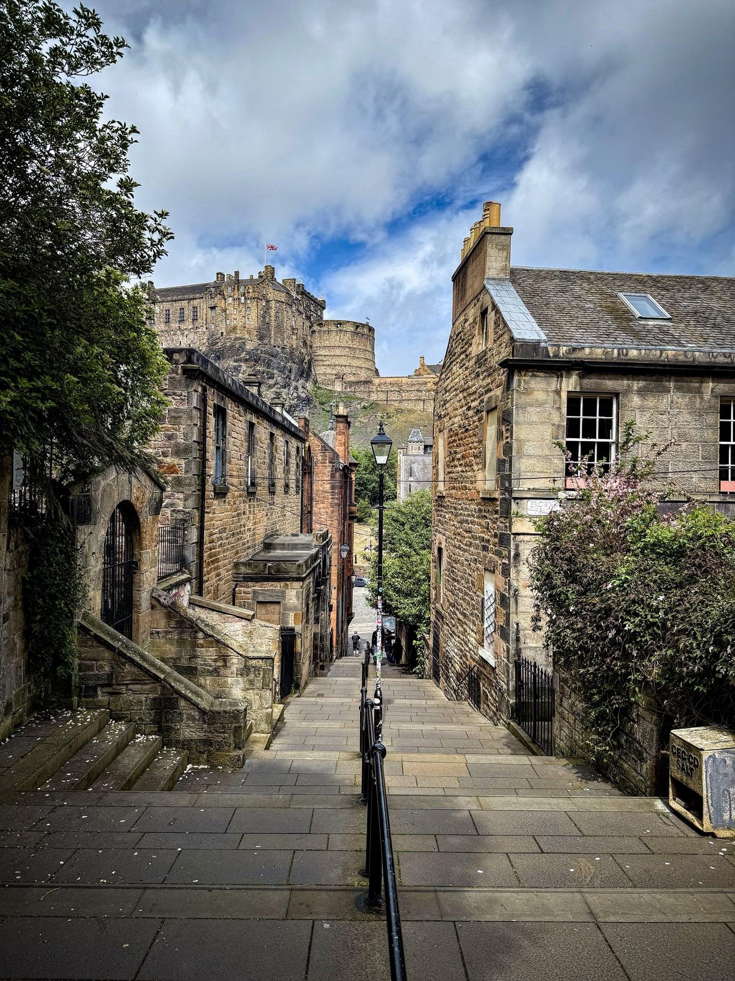 Narrow cobblestone street lined with historic stone buildings leading to a large castle on a hill under a cloudy sky. Green foliage on the left, metal railing along the descending path, and scattered leaves on the ground.