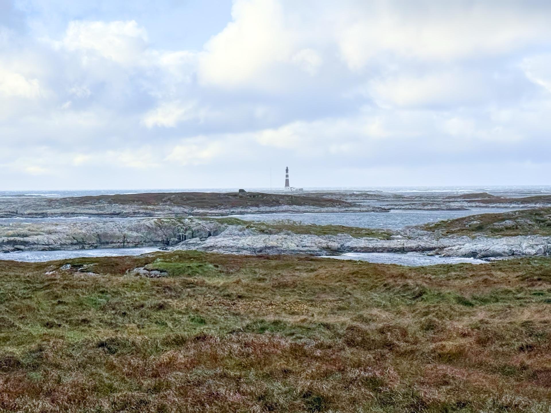 A coastal landscape featuring a white lighthouse in the distance, surrounded by flat rocky terrain with patches of grass. The foreground displays grassy fields transitioning to sparse rocky surfaces towards the sea under an overcast sky.