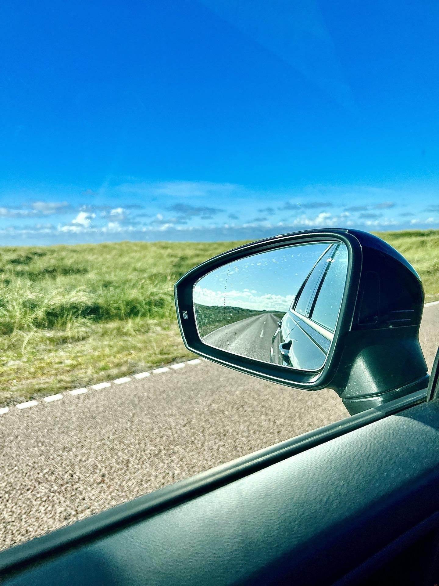 Side mirror of a car showing a reflection of a road extending into the distance; the outer scene includes a clear blue sky and a grassy field under bright sunlight.