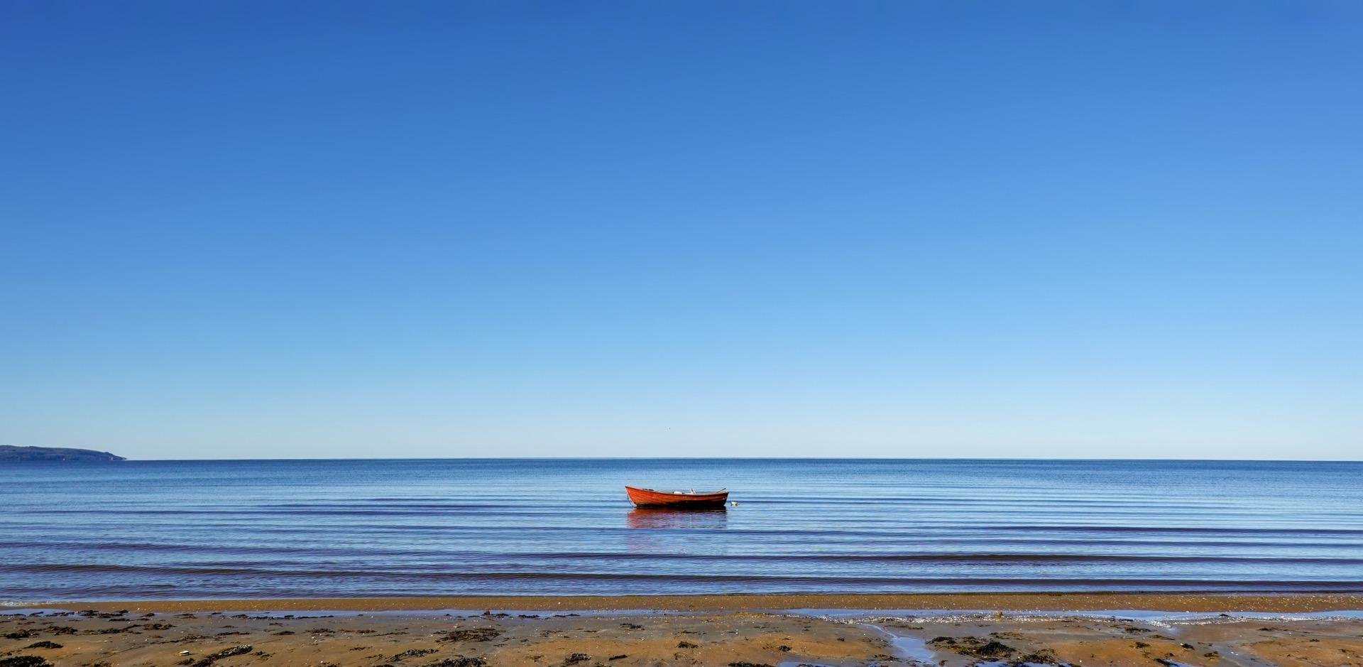 A lone orange boat floats on a calm sea under a clear blue sky, positioned centrally in the water. Visible in the foreground is a sandy shore with gentle waves lapping at the edge. The background features a faint outline of a coastline.