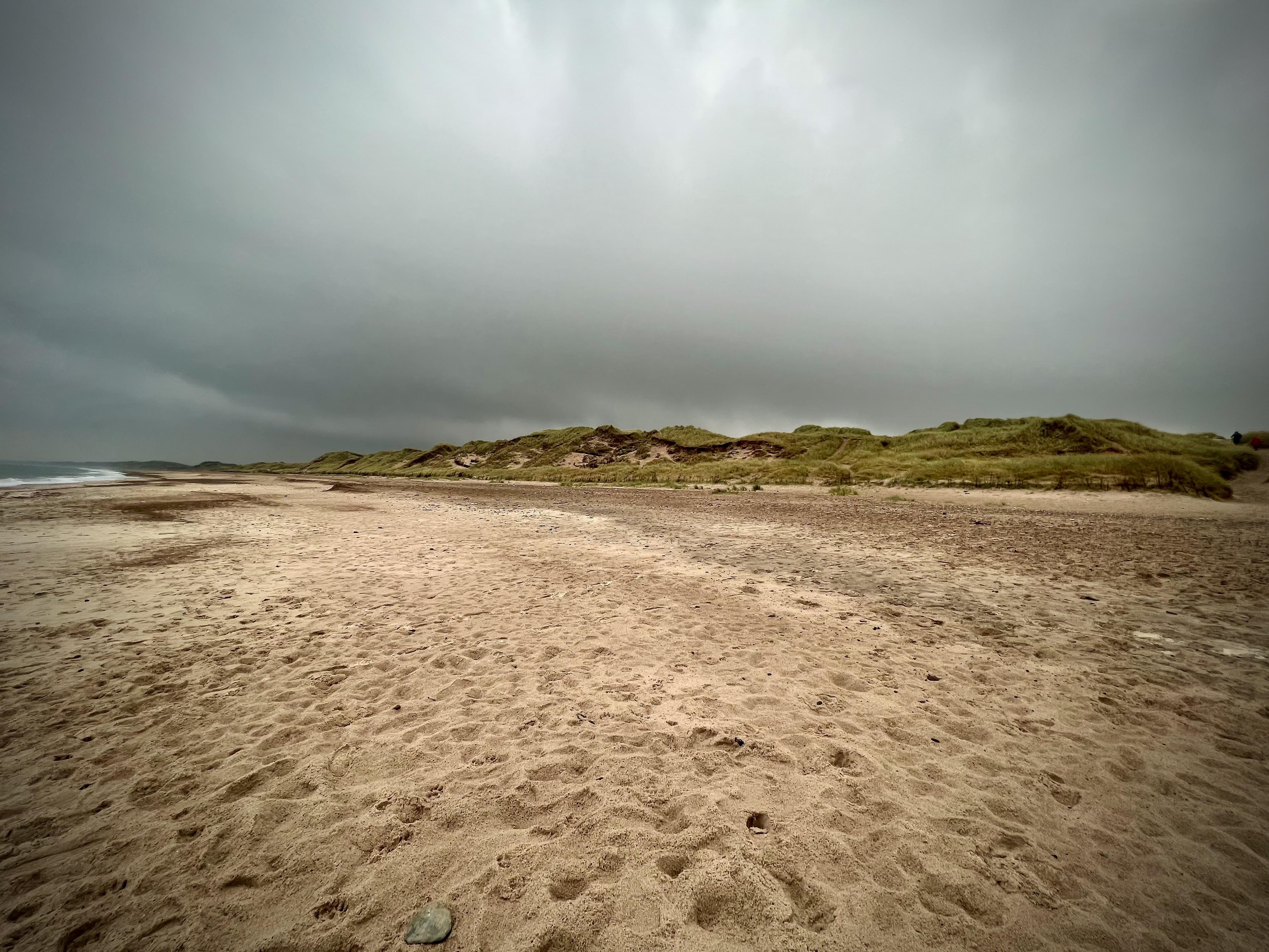 Wide sandy beach under an overcast sky with looming clouds, featuring textured sand in the foreground and rolling dunes covered with green grass in the background.