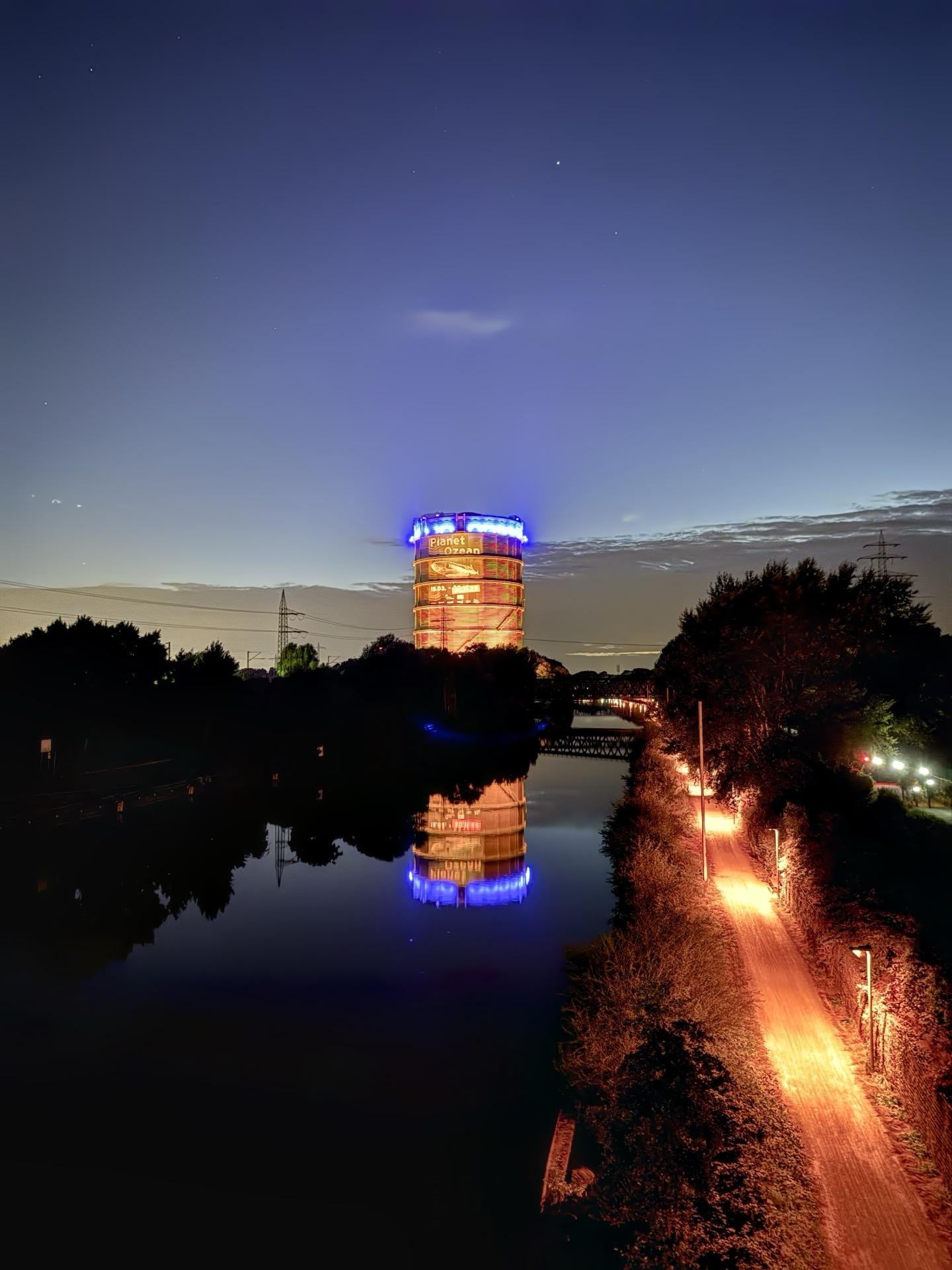 A nighttime view featuring a vertical, cylindrical tower illuminated with vibrant red and blue lights, reflected in the calm surface of a river. Alongside the river, a trail with soft orange lighting stretches into the distance, bordered by patches of foliage. The backdrop is a twilight sky with faint stars. Additionally, there is an electrical tower silhouette against the sky to the right.
