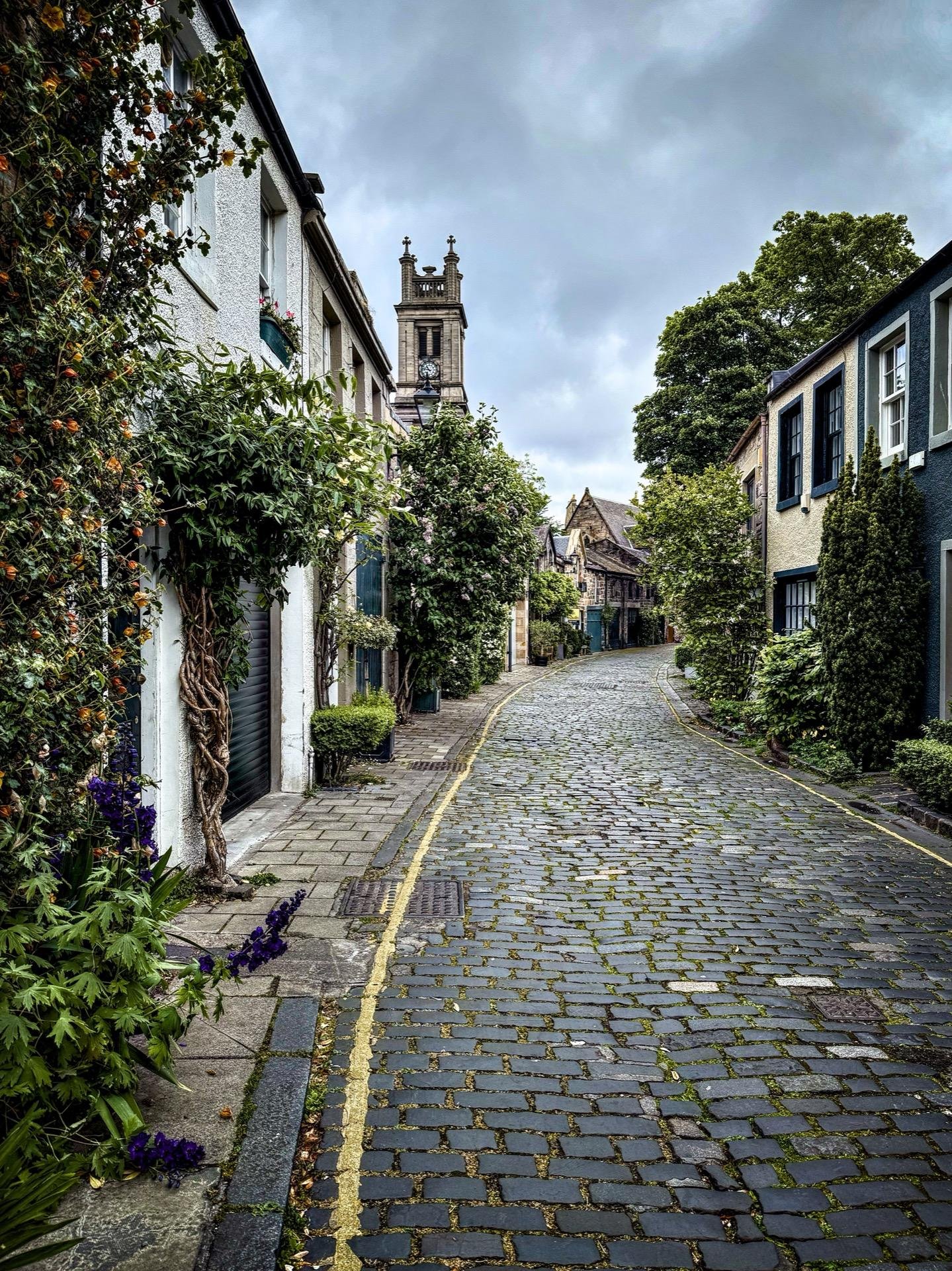 Cobbled street lined with colorful houses and abundant greenery under an overcast sky. A prominent church tower stands in the background. Flowers bloom along the sidewalk, adding touches of purple to the scene.