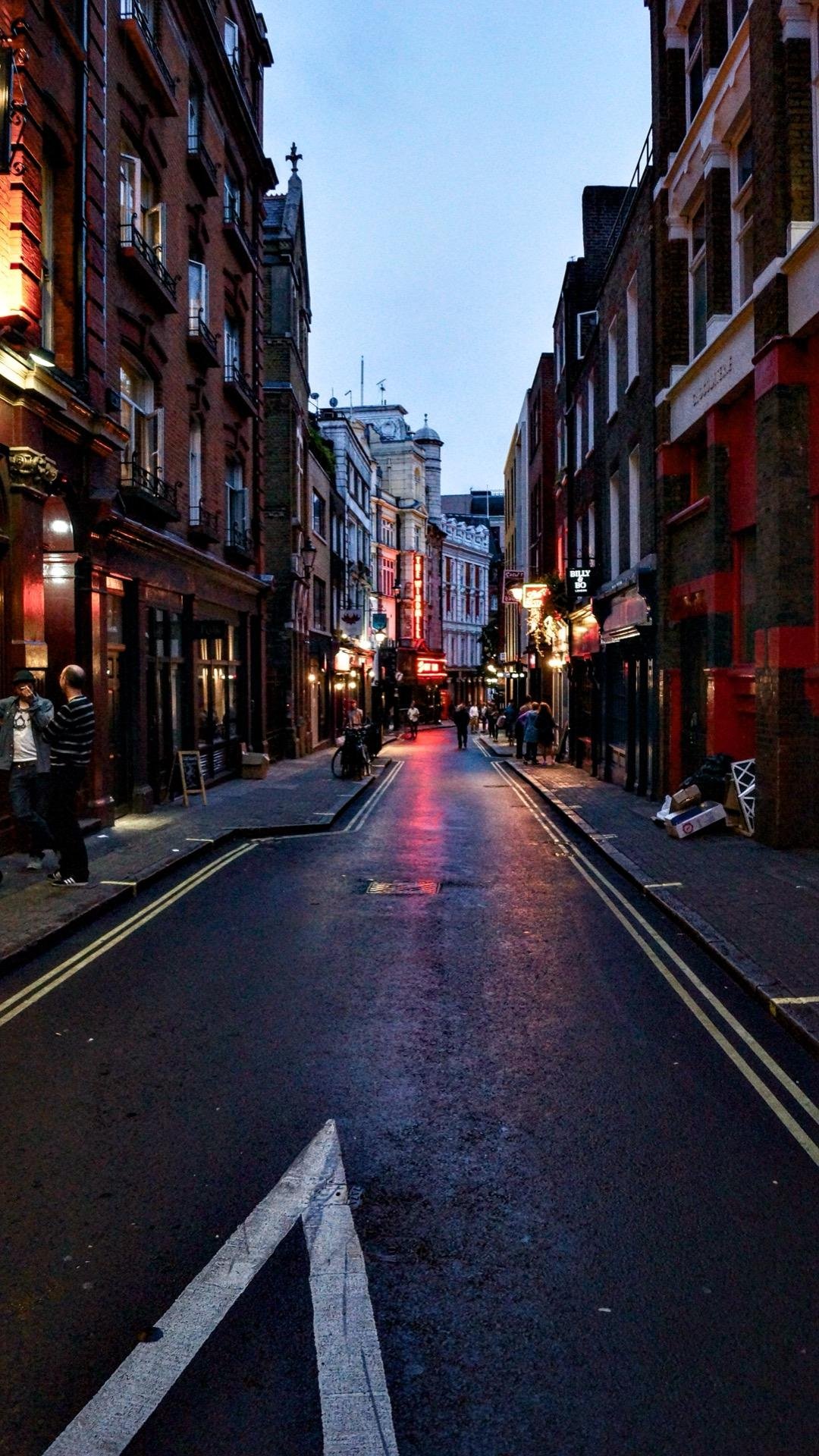 Narrow city street at dusk lined with old multi-story buildings illuminated by streetlights and neon signs. People walk along the sidewalks, and a wet road reflects the lights.