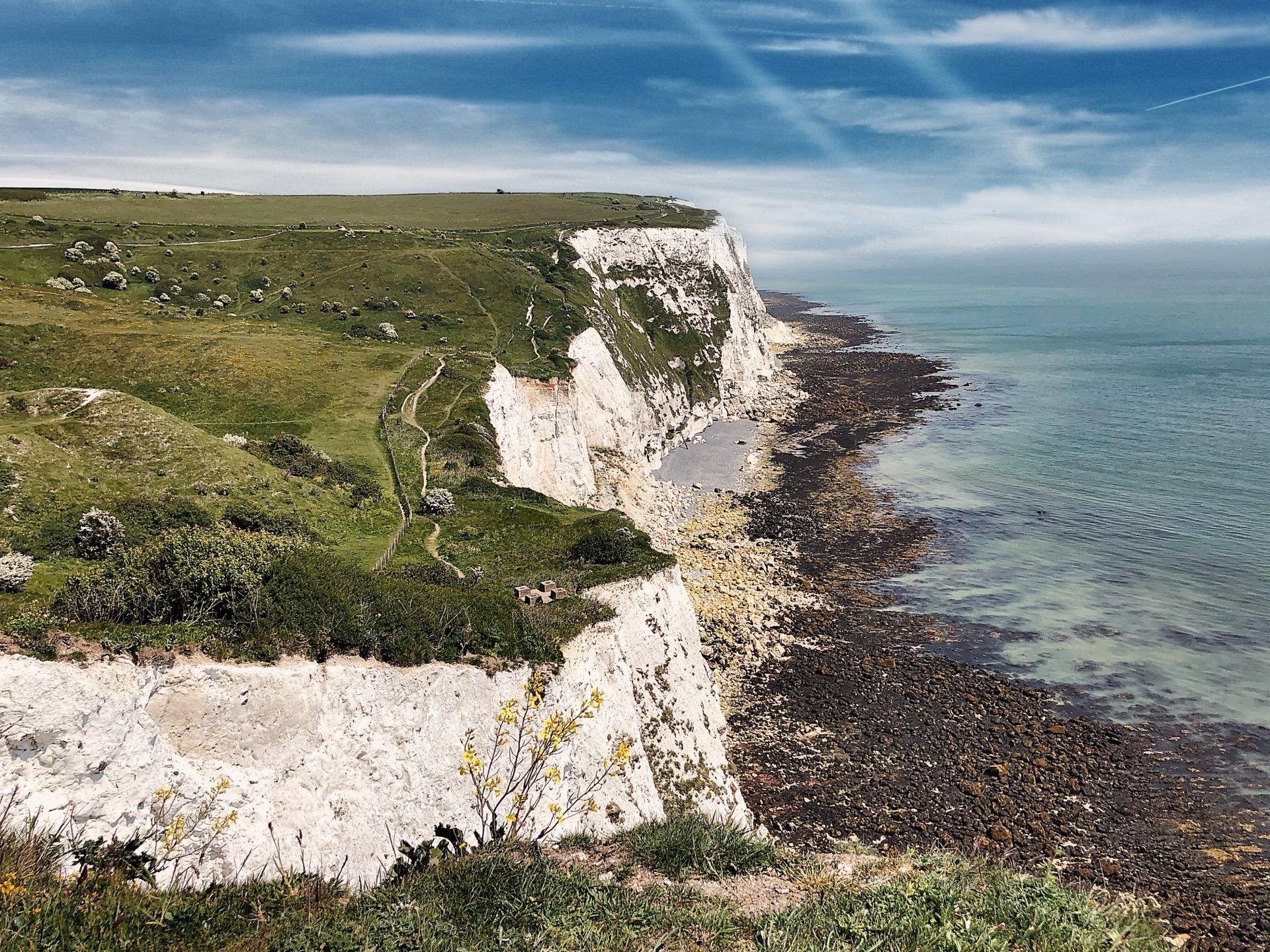 Lush green hillside with a winding footpath leading down to a white chalk cliff edge overlooking a clear blue sea. The coastline is adorned with rocky beach below, under a mostly clear sky with faint trails of clouds. Grazing sheep are visible on the distant hilltops.