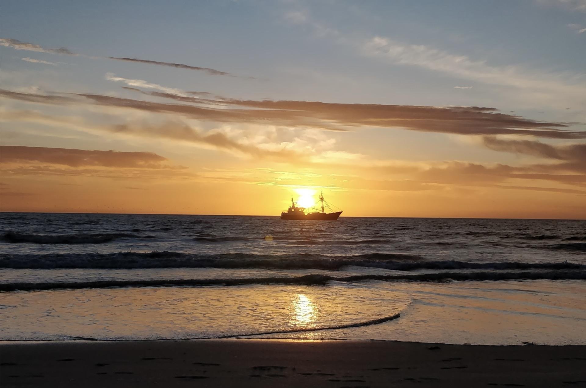 Sunset over the ocean with a fishing boat silhouetted against the bright sun, seen between partly cloudy skies and gentle waves approaching the sandy shore.
