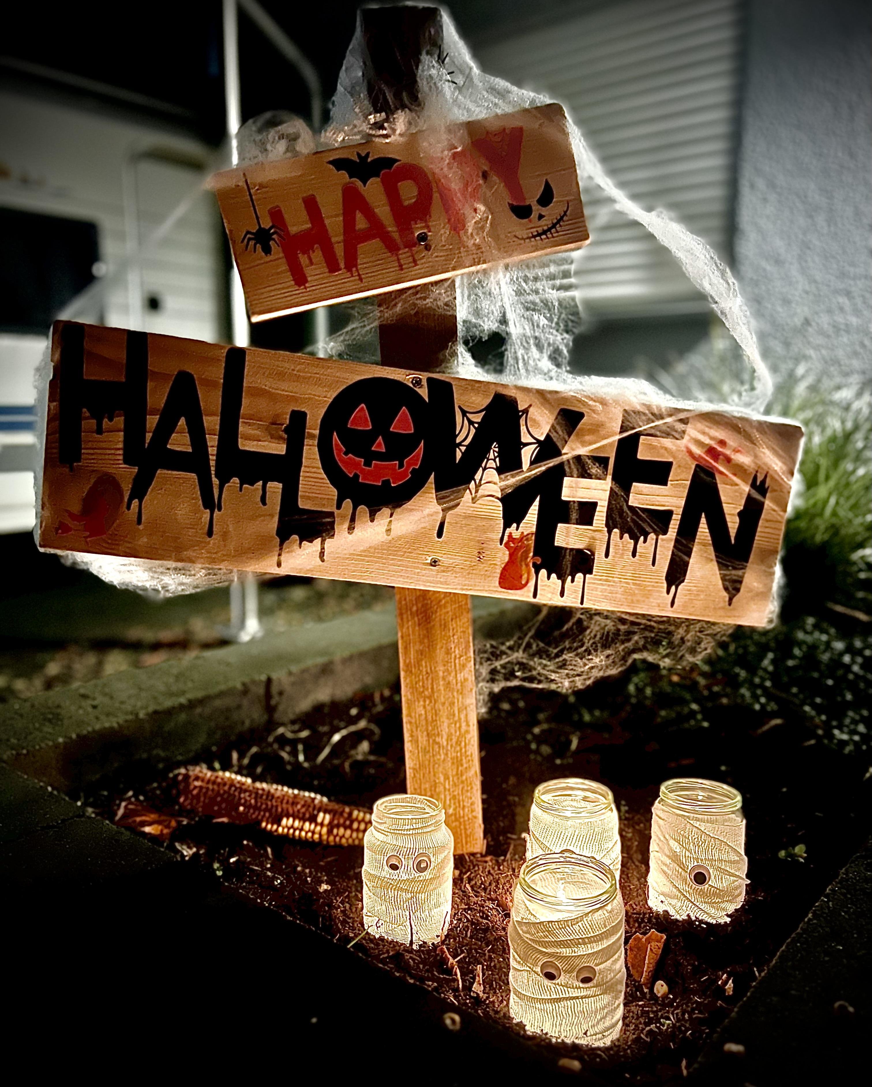 Wooden Halloween signpost decorated with cobwebs features two arrows; top reading "Happy!" with spider motifs and bottom "Halloween" with pumpkin and bats, illuminated by three candle lanterns wrapped in twine with cut-out owl faces, set against a night-time backdrop.