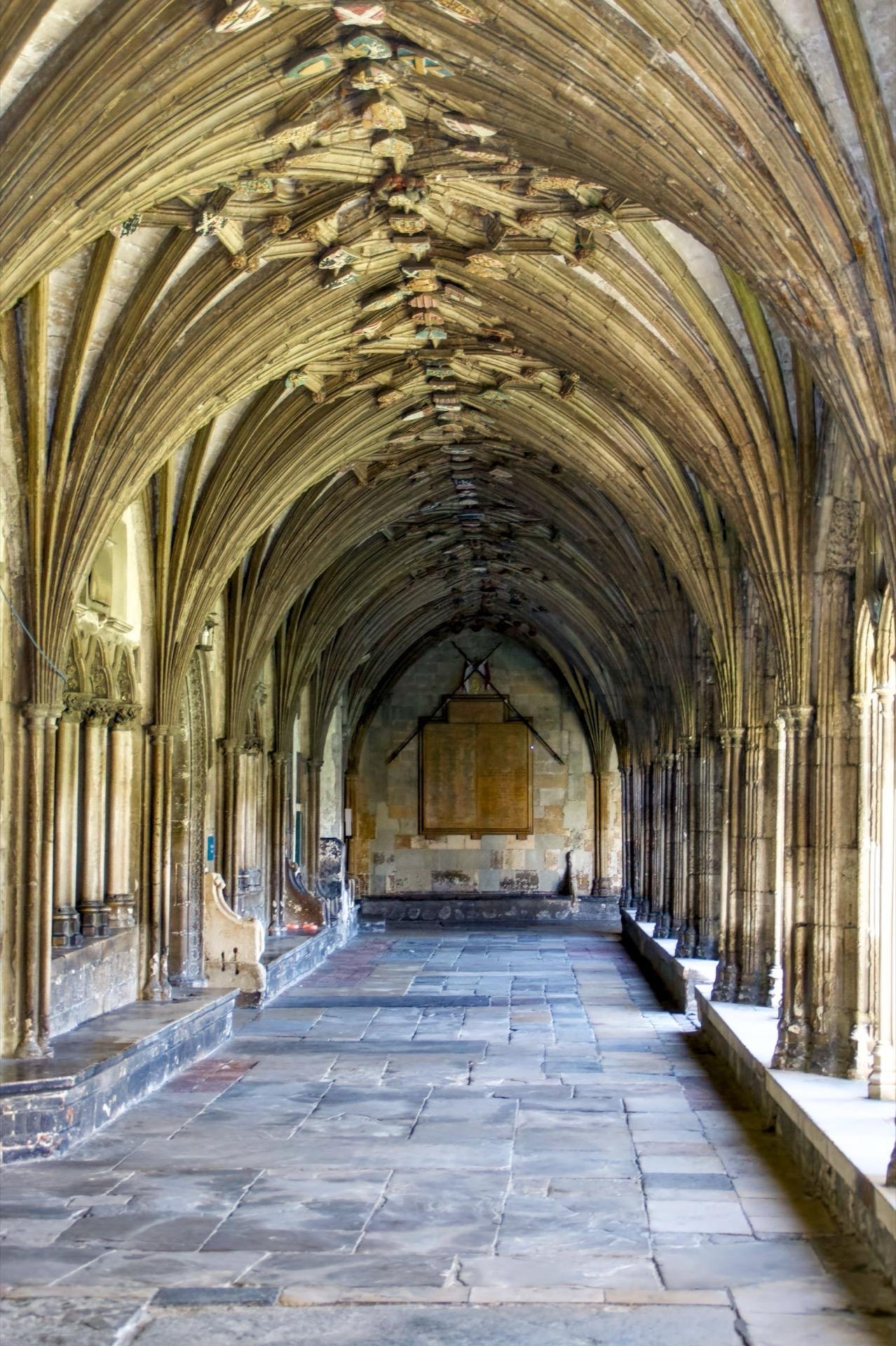 Vertical image of an ornate gothic cloister passage with vaulted stone arches and intricate ribbed ceilings. The pathway features worn gray stone slabs and is lined with numerous slim columns adorned with detailed carvings. Near the far end, an enclosed glass lantern hangs above a placard mounted on the wall. Sunlight filters through openings on the right, creating a play of light and shadows along the corridor.