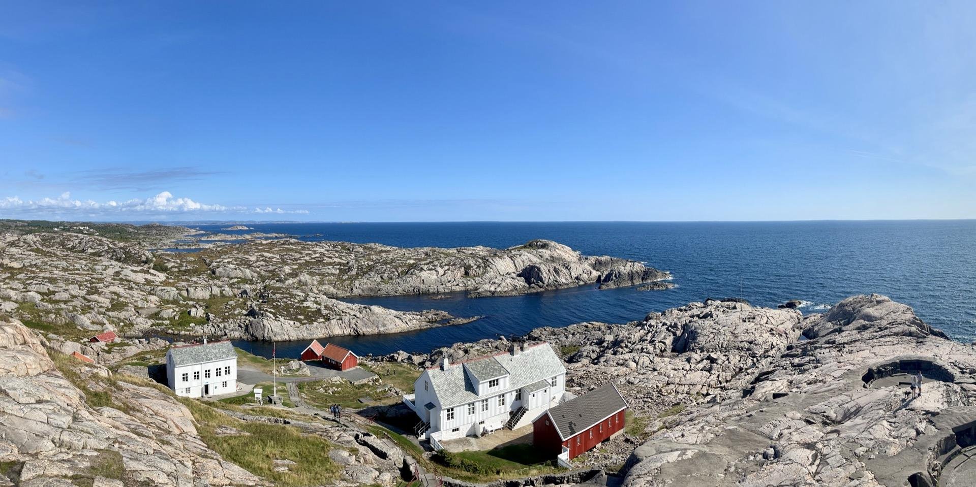 Panoramic view of a rugged coastal landscape featuring a clear blue sky, a calm sea, and a cluster of white and red buildings nestled among rocky terrain.