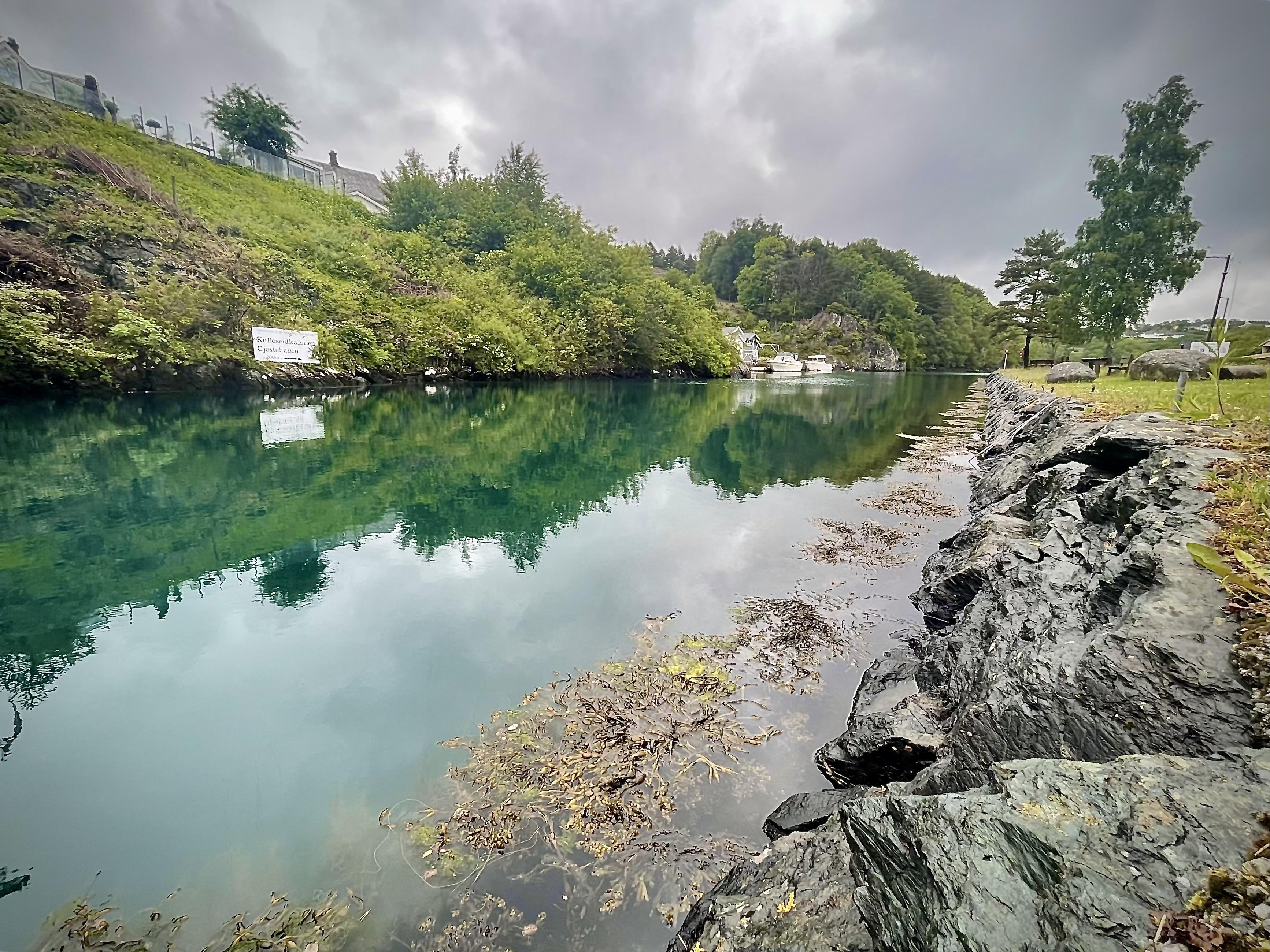 Calm lake with clear turquoise water reflecting a cloudy sky, bordered by rocky shores and lush greenery. A sign on the left bank and scattered floating debris near the foreground enhance the natural setting.