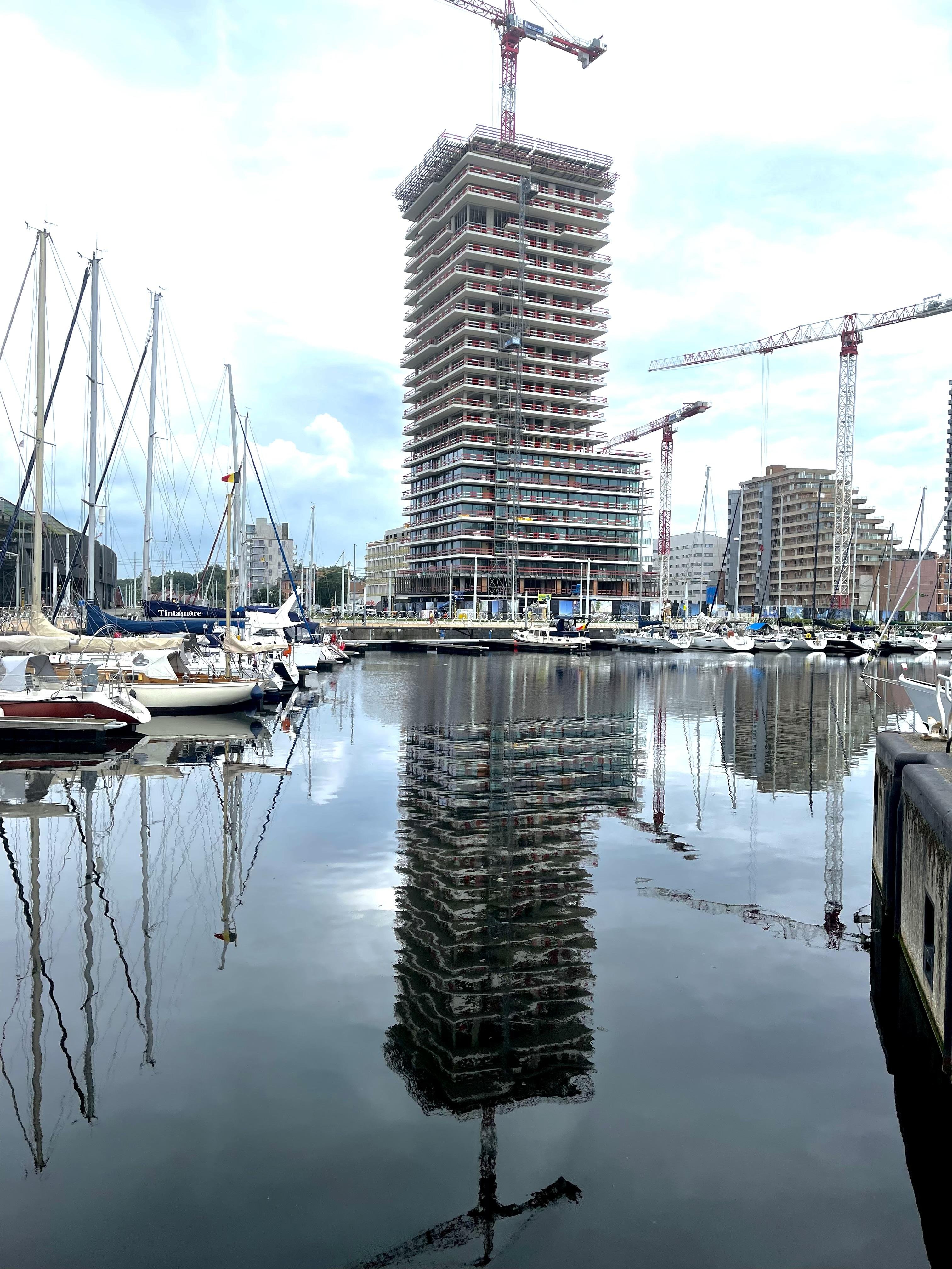 High-rise building under construction by a marina with multiple cranes surrounding it. A variety of sailboats are docked at the marina, and the building along with the masts of the boats are reflected in the calm water. The sky is overcast.