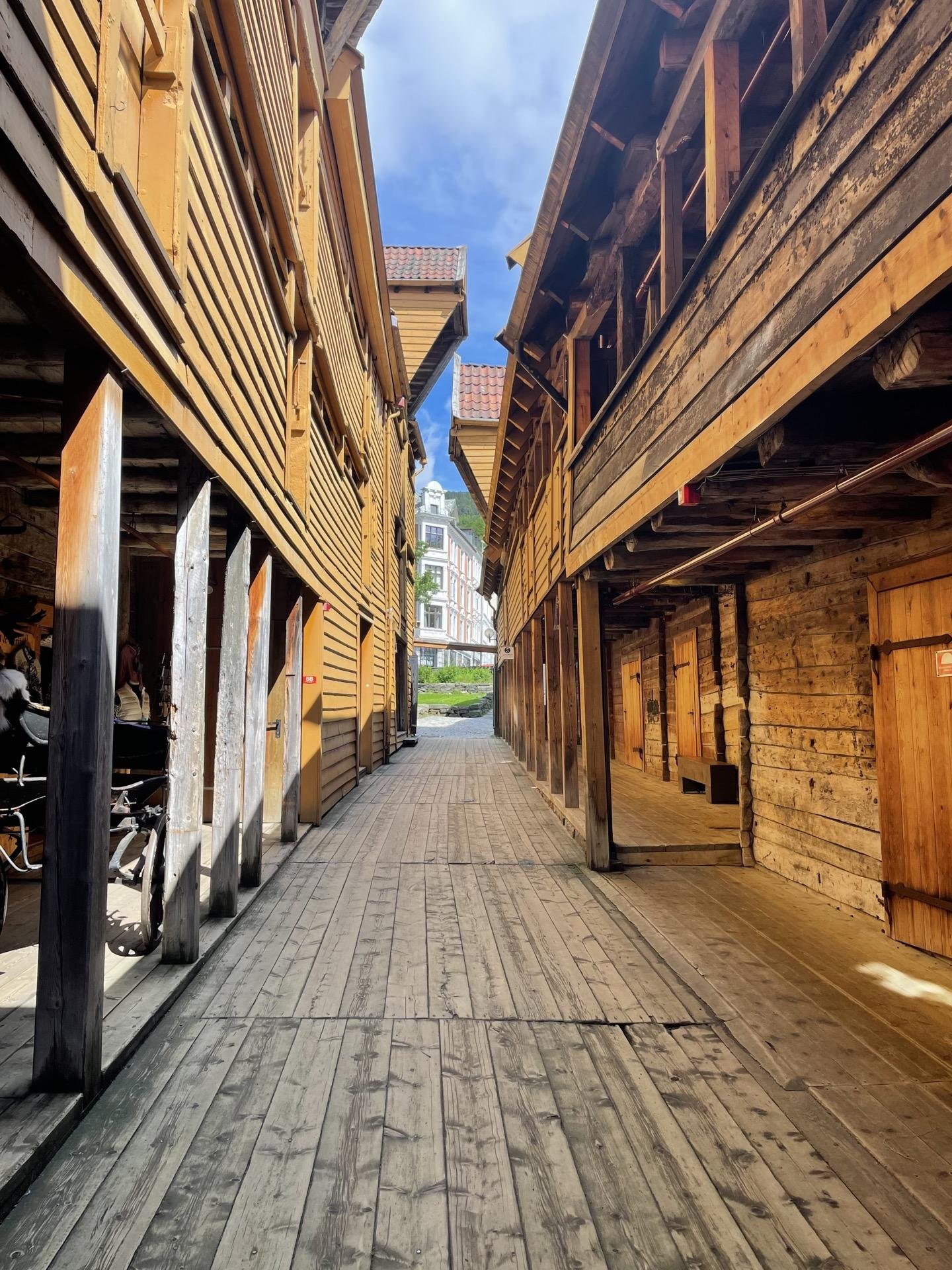 Narrow alleyway bounded by wooden buildings with high gables, weathered wood plank sidewalks, and a cobblestone street visible ahead. The architecture is rustic, featuring exposed beams and faded yellow paint. In the background, modern buildings with red rooftops are seen under a blue sky with few clouds.