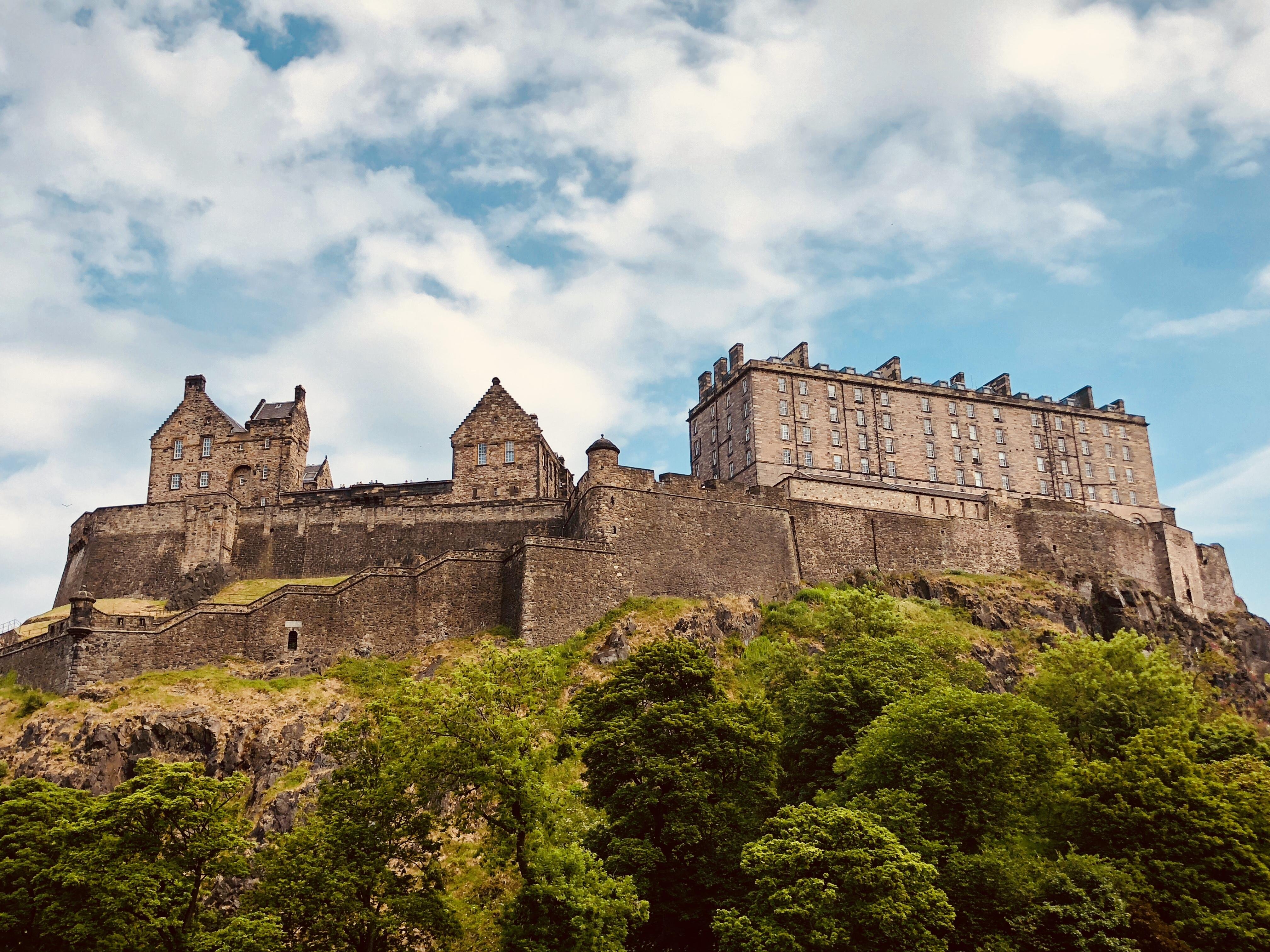 Ancient castle perched atop a large hill, surrounded by lush greenery, under a blue sky with wispy clouds. The stone structure features multiple towers and windows, with a rugged cliff base visible.