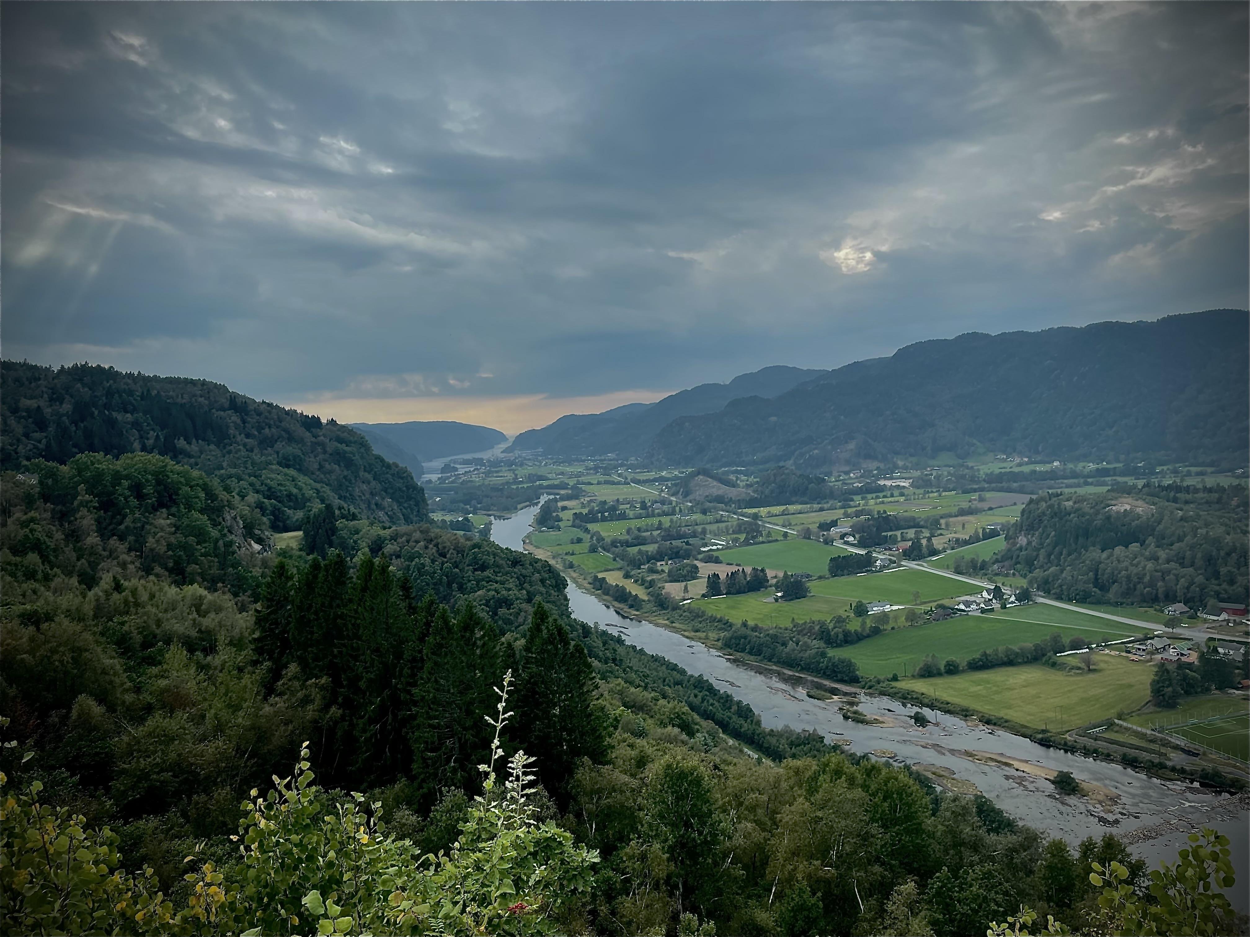 Landscape view of a river winding through a valley with lush greenery on either side, framed by forest-covered hills under a cloudy sky with visible sun rays piercing through on the left.