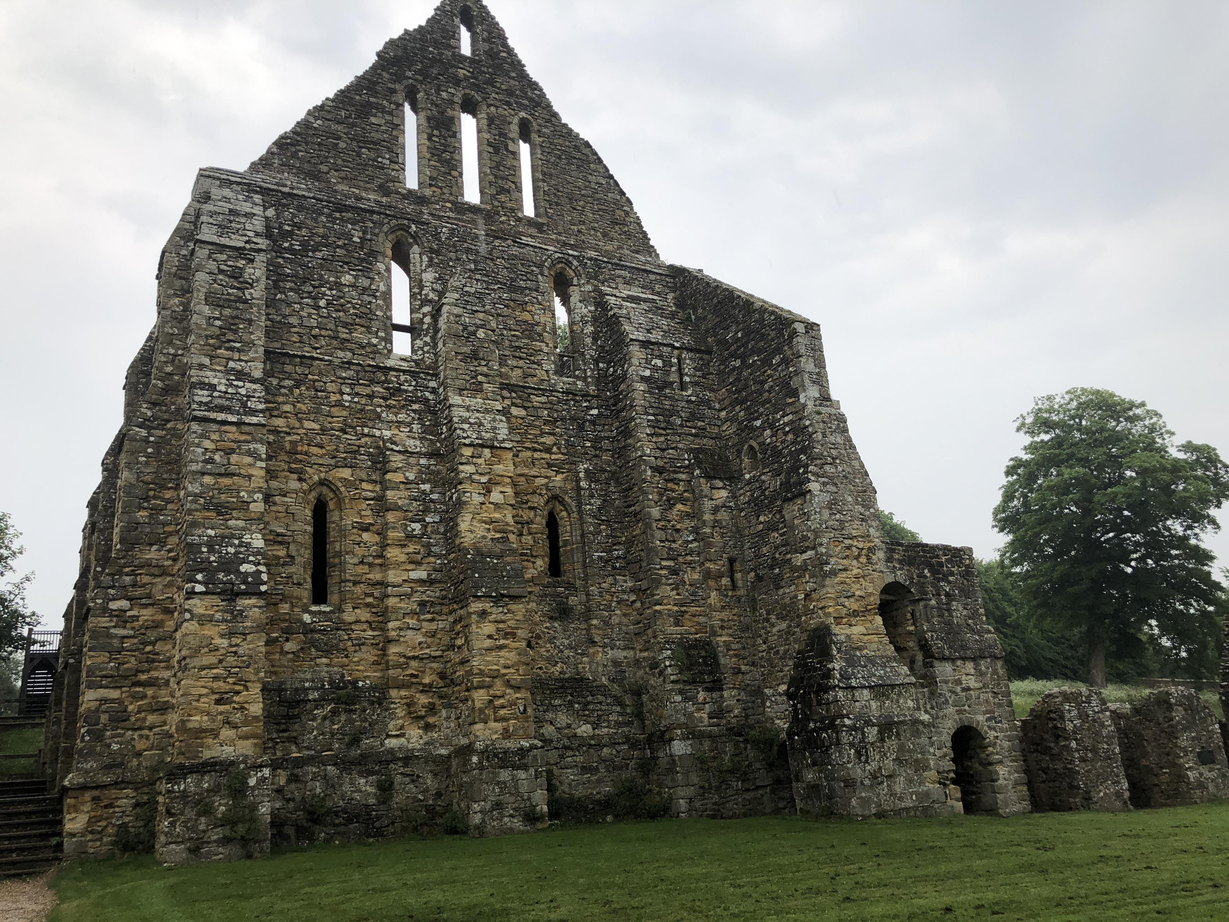 Ruined medieval stone abbey with several arched windows, viewed from the front, under a cloudy sky. The abbey has a large, triangular gable with three vertical openings at the top. Some greenery is visible at the base of the ruin, and trees can be seen in the background.