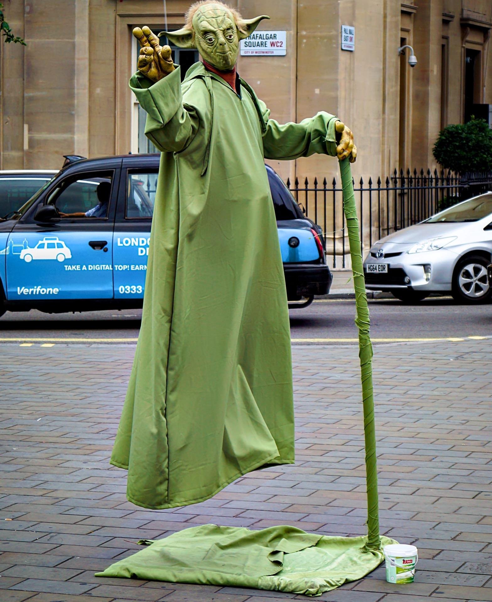 A street performer dressed as Yoda from Star Wars, appears to float with the aid of a green staff that extends to the ground covered in matching fabric. The performer is on a city street with passing cars and a blue taxi, near signage indicating Trafalgar Square. A tip jar sits on the pavement nearby.